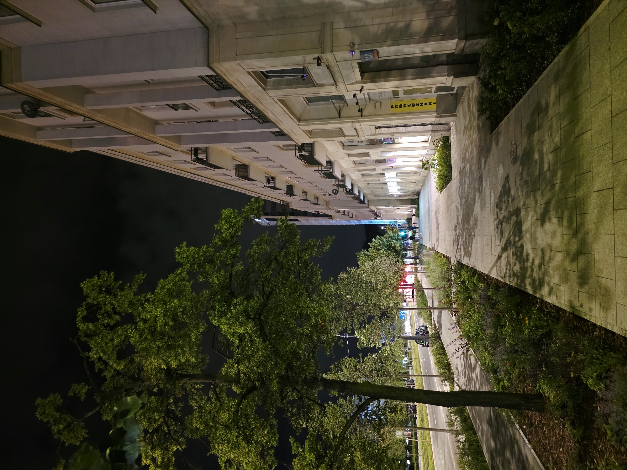 The image shows a nighttime street scene in Warsaw, Poland. On the right is a large, light-colored building with multiple floors and balconies. The building's facade is illuminated, highlighting architectural details and window frames. Below the balconies, there are ground-level businesses with illuminated signs. One prominent yellow sign reads "PODRECZNIKI." To the left of the building is a wide sidewalk paved with large gray stones. The sidewalk is lined with trees and greenery. Further to the left, there is a road with moving vehicles, including cars and a bus, visible under streetlights. A few people are walking on the sidewalk and a bicycle path is present between the sidewalk and the road. The sky is dark with some cloudy formations.