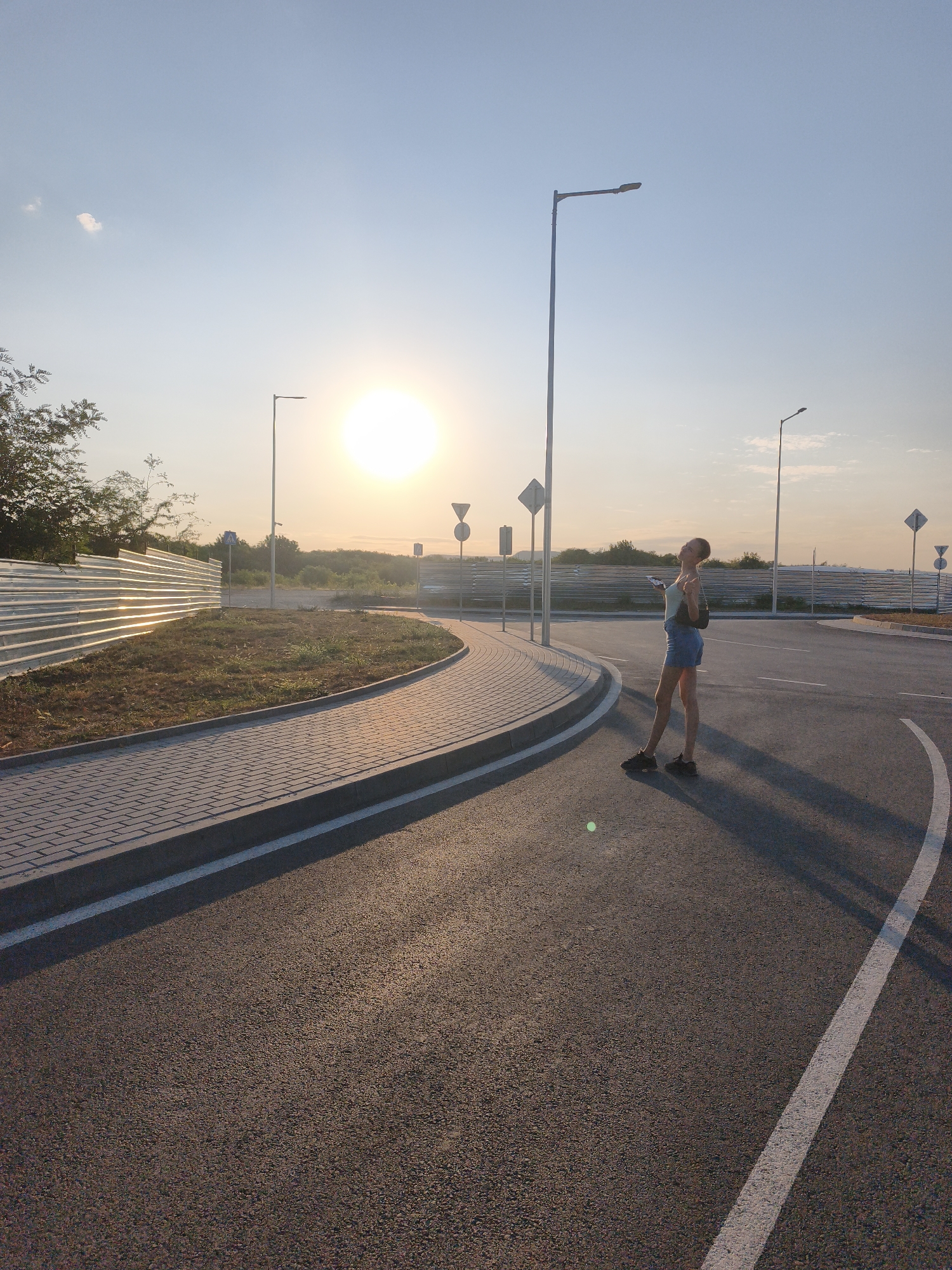 The image depicts a young woman standing on an asphalt road at what appears to be a roundabout. The sun is low in the sky, suggesting either sunrise or sunset, casting long shadows. The woman is wearing a light-colored top and denim shorts, with a dark bag slung over her shoulder. She is looking upwards and making a gesture with her hands. The road is lined with a gray brick sidewalk and a curved concrete curb. In the background, there is a metal fence, some vegetation, and several streetlights. The sky is a clear blue with a few wispy clouds. The scene is likely a newly developed area or a construction site given the appearance of the fence and road markings. There is no specific visual information in the image that would allow for the precise identification of the location as Lovech, Bulgaria.