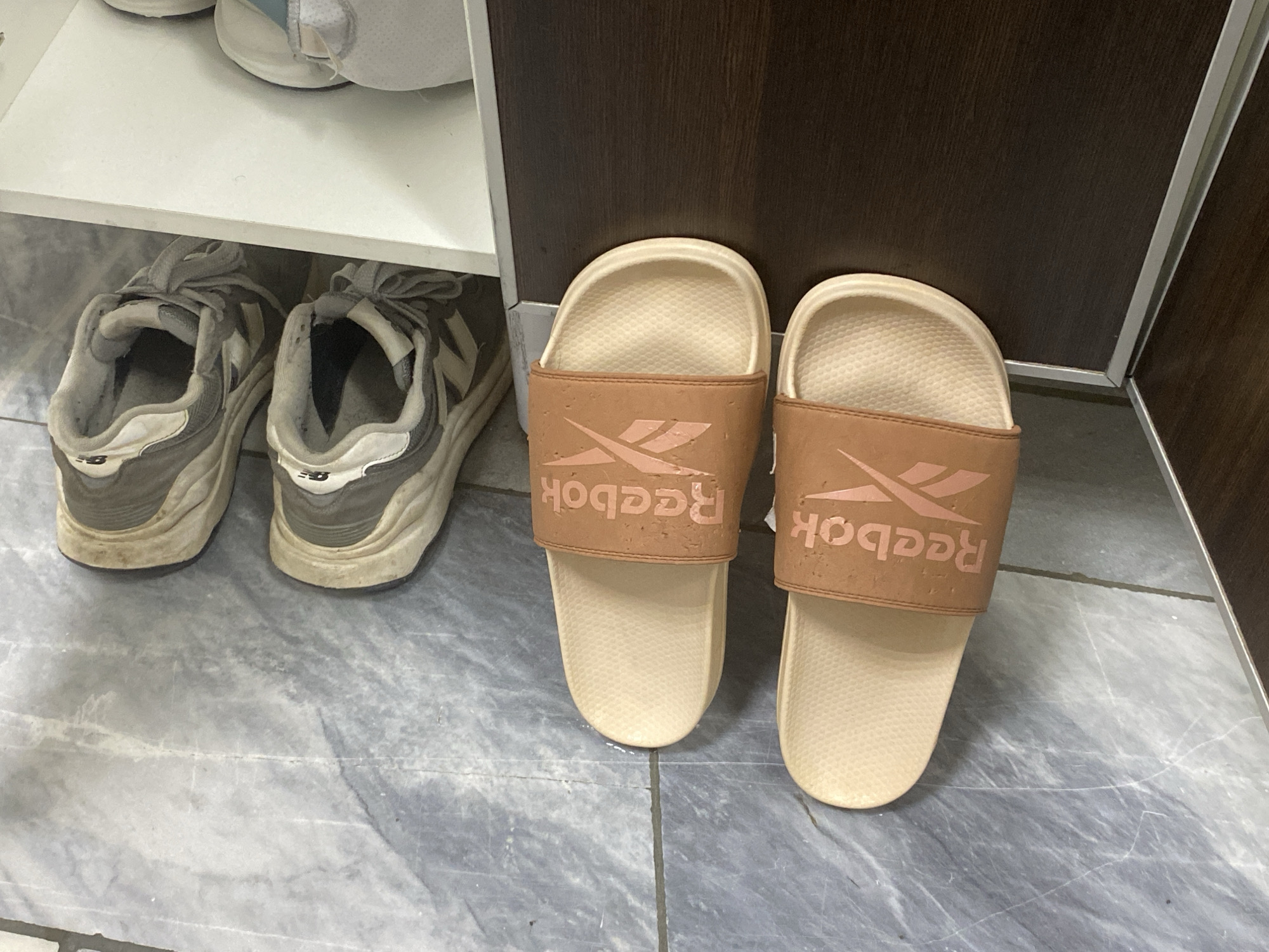 The image shows a pair of tan Reebok slides with a peach-colored Reebok logo on a textured, light-colored sole. They are positioned on a gray tiled floor in front of a dark brown wooden cabinet. To the left of the slides are a pair of gray and white New Balance sneakers, also on the tiled floor. The sneakers appear to be slightly worn. Above the sneakers is a white shelf, with another white shoe visible on it.
