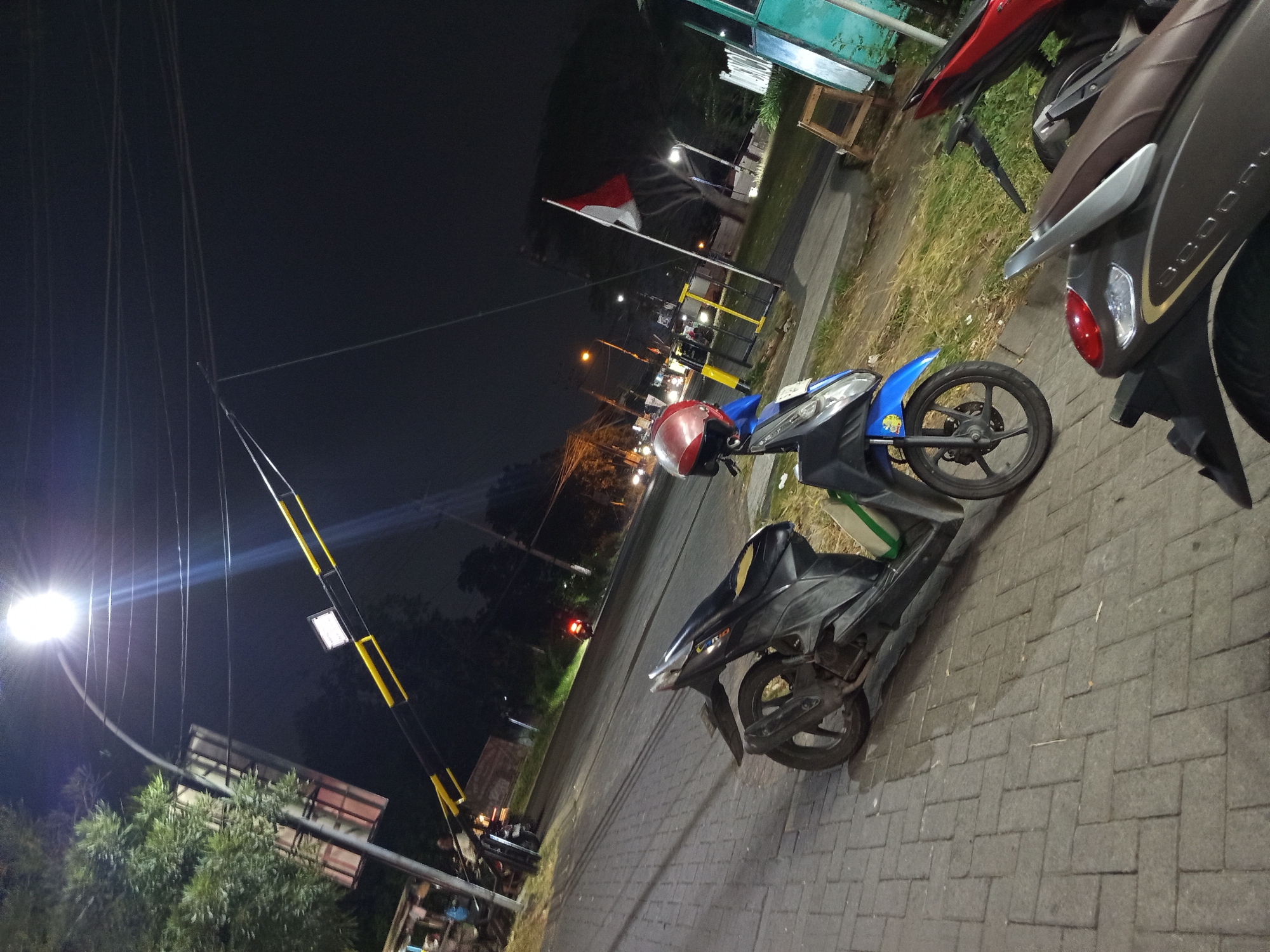 A dark, low-angle, nighttime outdoor shot captures a street scene in Gemurung, Indonesia, illuminated by a bright overhead streetlamp. The foreground features a black and blue automatic scooter parked on a brick-paved sidewalk. A red and silver helmet rests on the scooter's handlebars. Beyond the scooter, the street extends, with a yellow and black barrier arm crossing overhead, suggesting a checkpoint or entrance. A red and white Indonesian flag is visible on a flagpole to the right, slightly obscured by trees and darkness. In the background, the streetlights of passing vehicles are visible, and the silhouette of trees lines the far side of the road. The overall environment appears to be a moderately busy street at night.