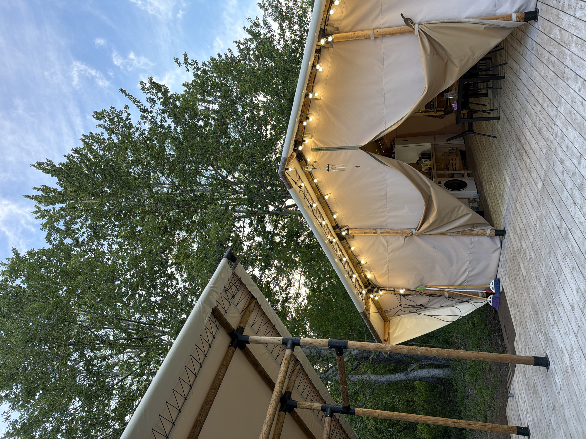 This media file captures an outdoor scene in Esso, Russia, likely during a pleasant evening. The focus is on two canvas tents, possibly part of a glamping setup. The larger tent, illuminated by strings of warm fairy lights along its edge, is partially open, revealing a glimpse of furniture and appliances inside, suggesting a functional living space. A wooden deck surrounds the tent, adding to the rustic charm. In the foreground, a smaller tent structure, supported by bamboo poles, is visible. The background is dominated by the lush green foliage of trees, indicating a natural, possibly forested, setting. The sky above is a mix of blue and wispy clouds, hinting at fair weather. The overall mood is serene and inviting, evoking a sense of outdoor relaxation and comfort. No people are visible in the frame. There is no visible text in the image.