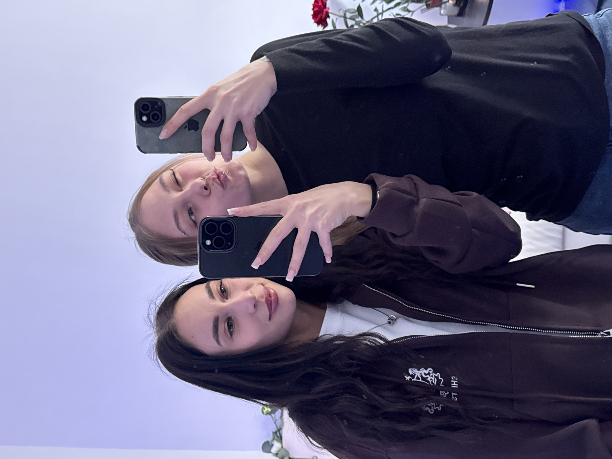 The image is a mirror selfie featuring two young women indoors. The subjects are positioned horizontally within the frame, with their heads on the left side.
The woman at the top has lighter, possibly blonde or light brown hair, and light-colored eyes. She is making a playful kissing face. She wears a black long-sleeved top and has a nose piercing and an ear piercing. She holds a dark green/grey iPhone with a triple camera module and visible Apple logo, appearing to be taking the selfie.
The woman at the bottom has dark brown, wavy hair and dark eyes. She has a slight, calm smile. She wears a dark brown zip-up hoodie over a white t-shirt. She is also holding a black iPhone with a triple camera module.
Both women have manicured nails with white tips.
In the background, a light-colored, possibly pale lavender wall or surface is visible. Towards the upper right of the frame, a red rose and some green foliage are discernible, suggesting a decorative plant.
The activity is the two women taking mirror selfies together. The overall mood conveyed is playful and relaxed.
Visible text on the dark brown hoodie of the bottom woman appears as mirrored white text and abstract designs, which, if un-mirrored, might spell "SHI ZT" or a similar brand/design.