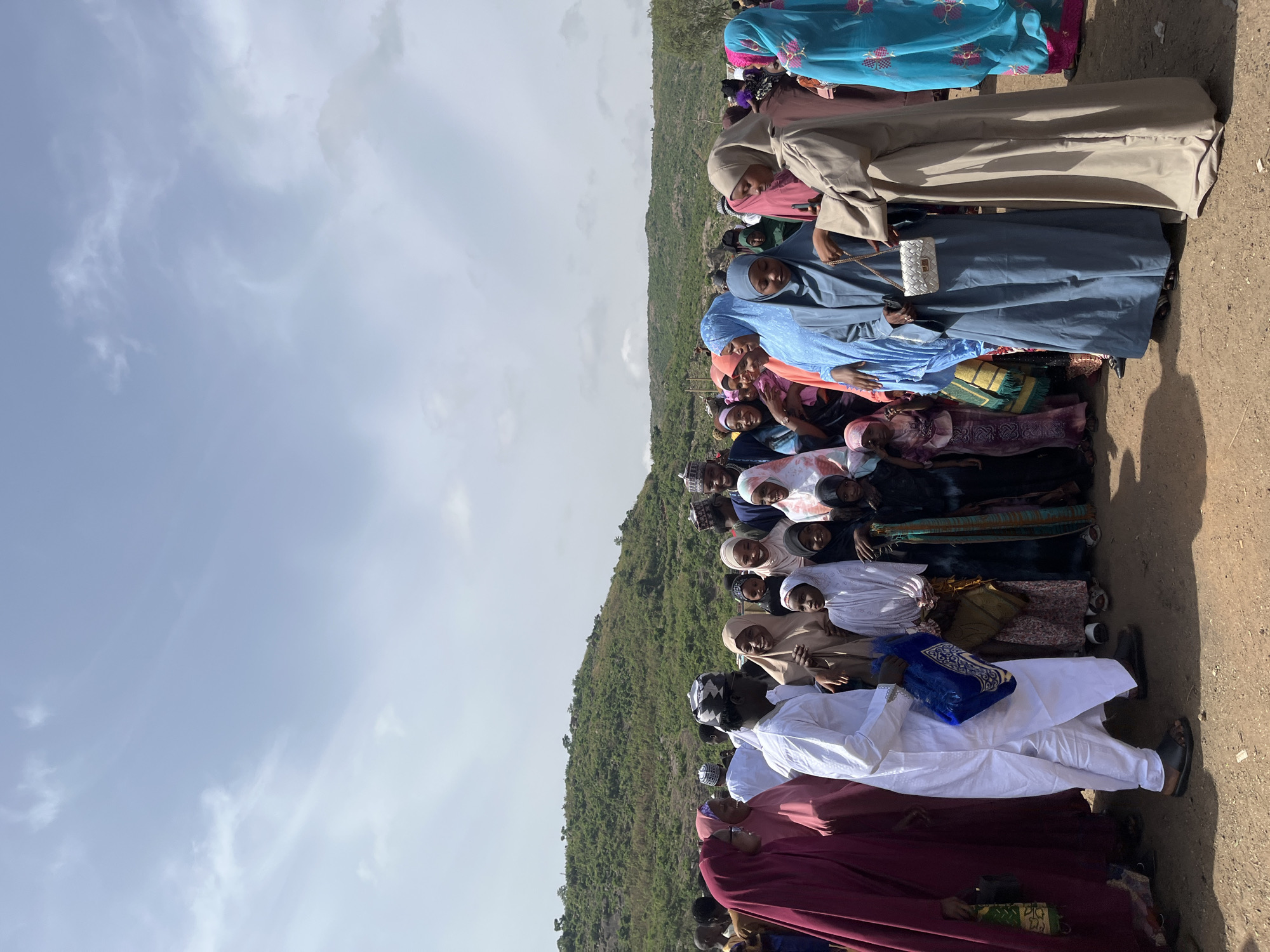 The image depicts a group of people lined up outdoors in Naraguta Hausawa, Nigeria. Most of the people are wearing head coverings, possibly hijabs, and traditional clothing. A hillside is visible behind them, and the sky is visible above. The ground is dirt. The people appear to be standing or sitting close together. Some individuals are carrying bags or other personal items.
