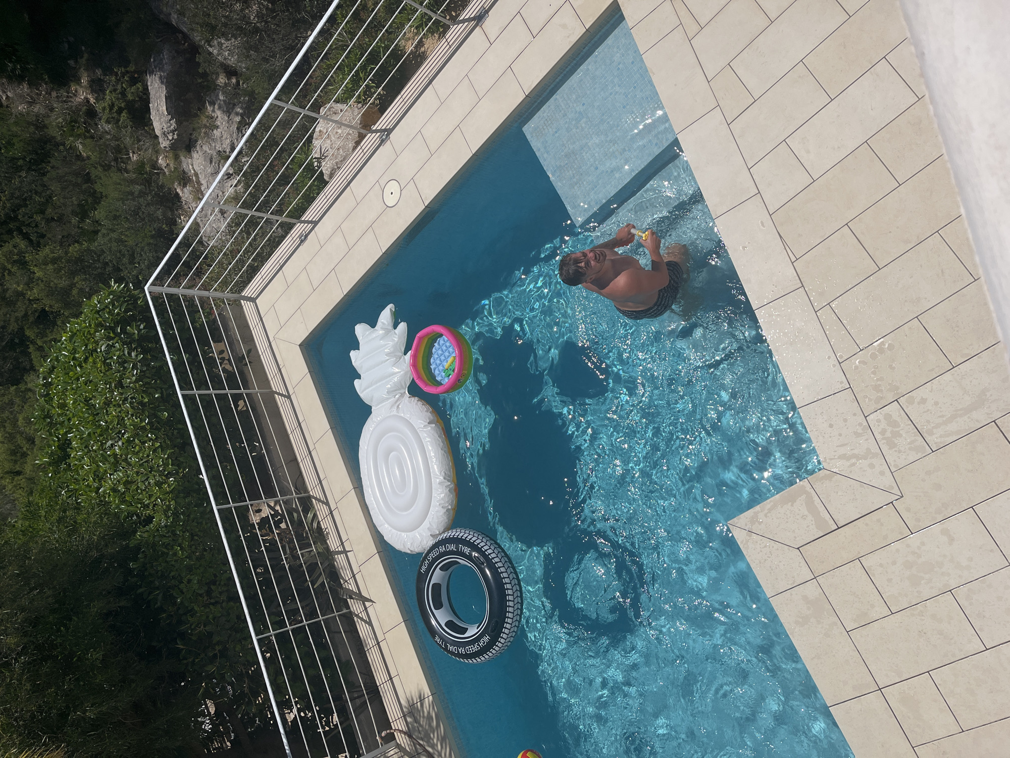 The image shows a backyard swimming pool on a sunny day in El Poble Nou de Benitatxell/Benitachell, Spain. The pool is surrounded by light-colored rectangular tiles. A white metal railing runs along the edge of the property bordering a green area with trees and rocks. Inside the pool, a man in swim trunks is holding a yellow water gun. Several inflatable pool toys are also floating in the water: a white pineapple, a rainbow-colored circle with small balls, and a black tire. The water is clear and blue, reflecting sunlight.