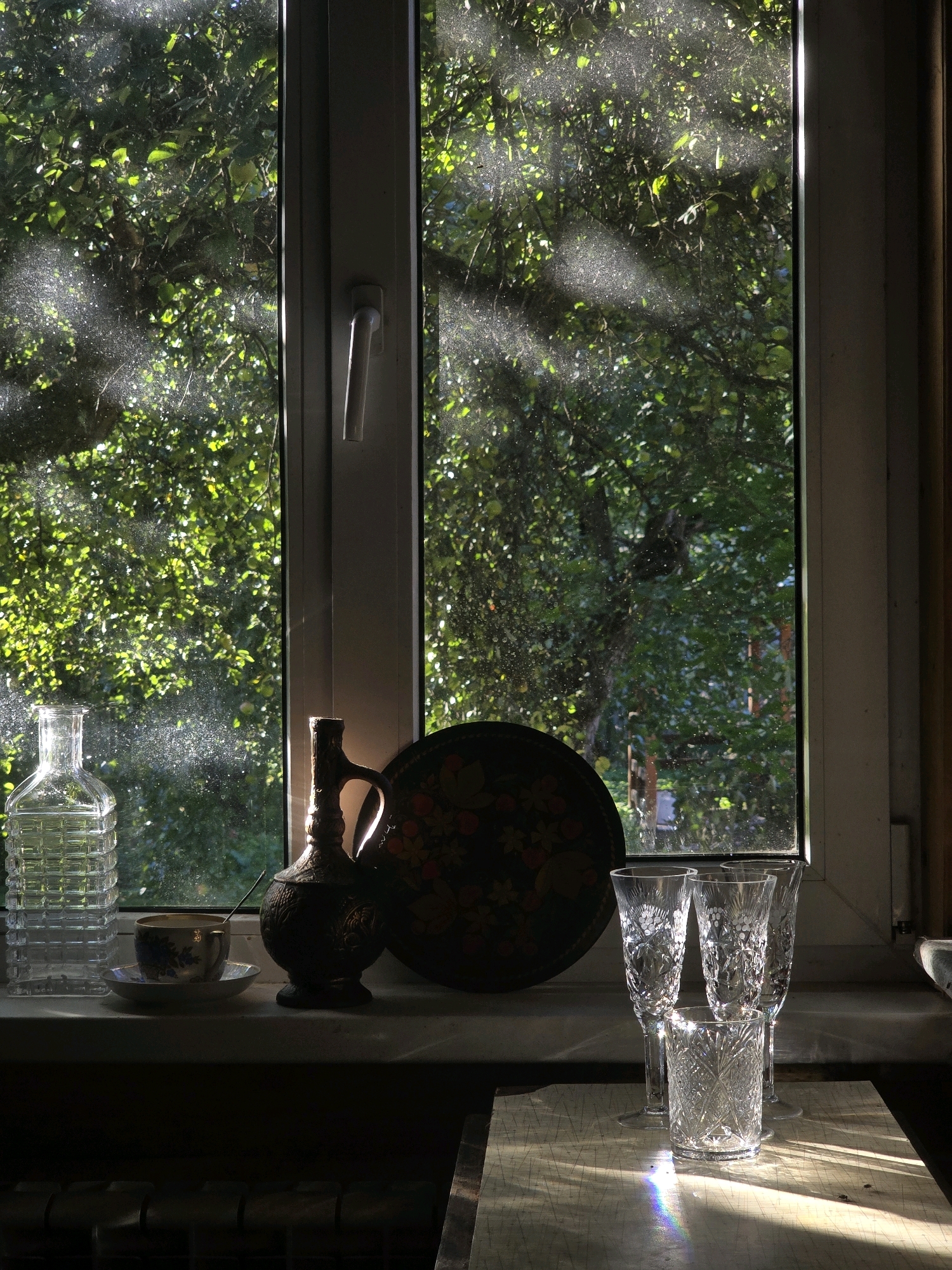 The image depicts a still life on a windowsill overlooking lush green foliage. The scene is illuminated by dappled sunlight filtering through the leaves and window. On the windowsill are several objects: a clear glass decanter with a textured surface on the left, a small teacup and saucer with a blue floral pattern in the center, and a dark, ornate jug to the right of the teacup. Behind these items, a large, dark decorative plate with a floral design is visible. Further to the right, three crystal glasses are arranged: two tall, slender champagne flutes and one shorter, wider tumbler. The background through the window shows dense green trees and leaves, with light glinting off dust particles on the glass. The windowsill itself appears to be made of a light-colored material, and the area below the windowsill is dark and out of focus, suggesting the presence of a radiator or heating element. There are no people or animals present in the image. The overall impression is one of a quiet, sunlit domestic interior.