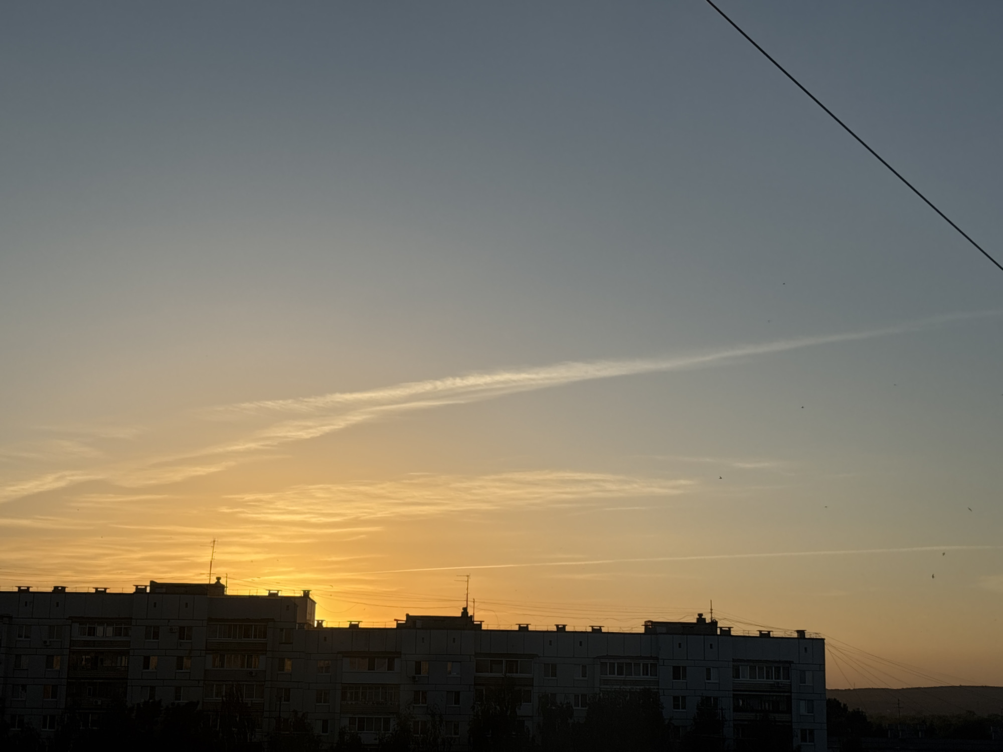 The image depicts an urban scene during twilight or sunrise. In the foreground, a large, multi-story residential building with numerous windows and balconies dominates the view. The building's silhouette is partially illuminated by the warm light of the rising or setting sun. Above the building, the sky transitions from a pale yellow-orange near the horizon to a lighter blue-gray higher up. There is a thin contrail visible in the sky, possibly from an airplane. A power line stretches across the upper right corner of the frame. The presence of the building suggests the location is in a city, but the specific city is Unknown. The silhouette of trees and possibly hills can be seen in the far background, further suggesting the edge of an urban area. No people are visible in the image.
