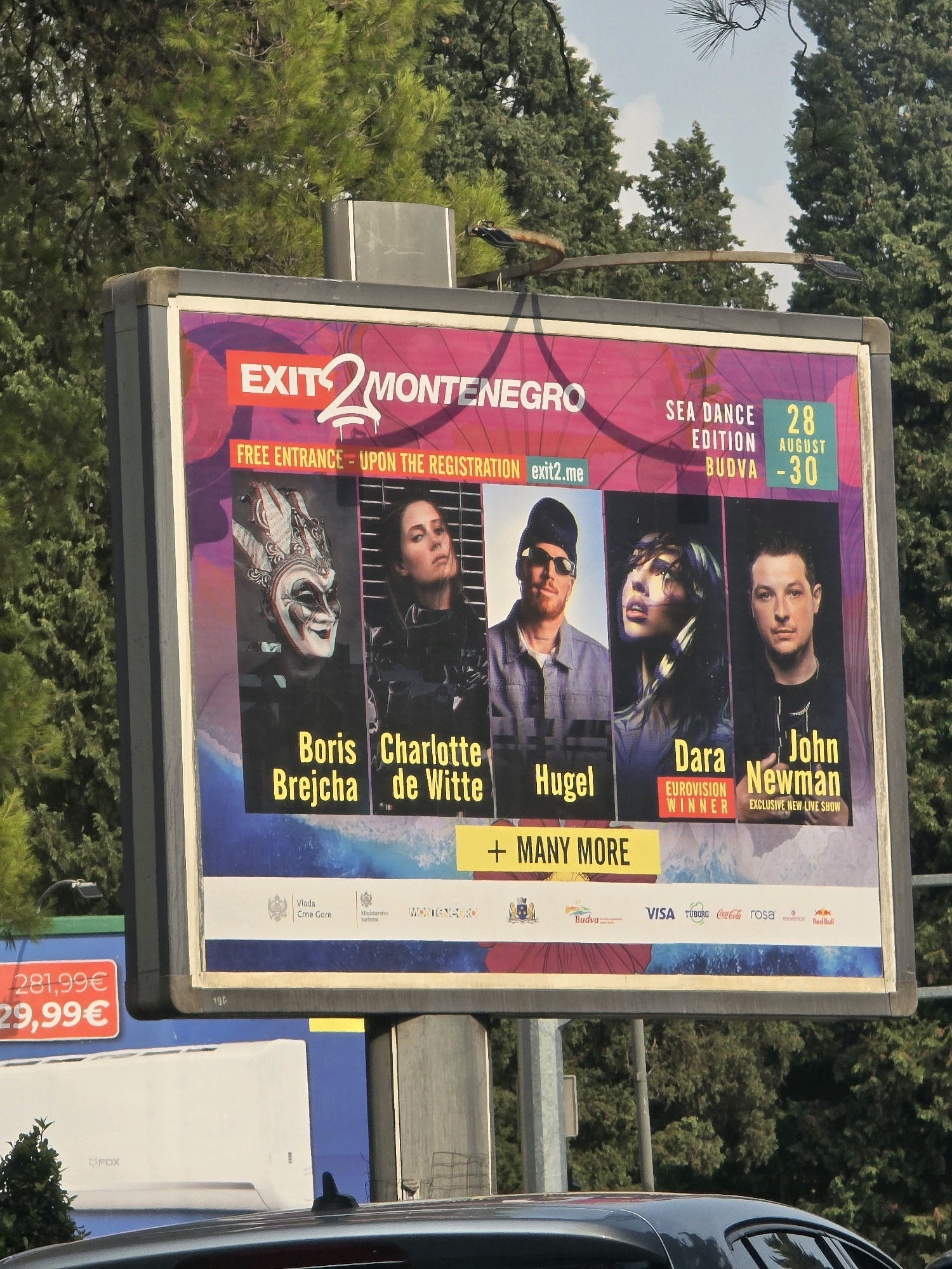 A large outdoor billboard is displayed in Podgorica, Montenegro, advertising the "EXIT 2 MONTENEGRO" festival. The billboard features images of performers, including Boris Brejcha, Charlotte de Witte, Hugel, Dara, and John Newman. It also includes event details such as "SEA DANCE EDITION BUDVA 28 AUGUST - 30" and "FREE ENTRANCE - UPON THE REGISTRATION exit2.me". The billboard is positioned against a backdrop of lush green trees and a partly cloudy sky. Below the main advertisement, various sponsor logos are visible, including Visa, Tuborg, Coca-Cola, Rosa, Essence, and Red Bull. In the foreground, the top of a dark-colored car is visible, suggesting the billboard is located near a road. To the left of the main billboard, a smaller blue advertisement with red text displaying "281,99€" and "29,99€" is partially visible. The scene appears to be set during daylight hours.