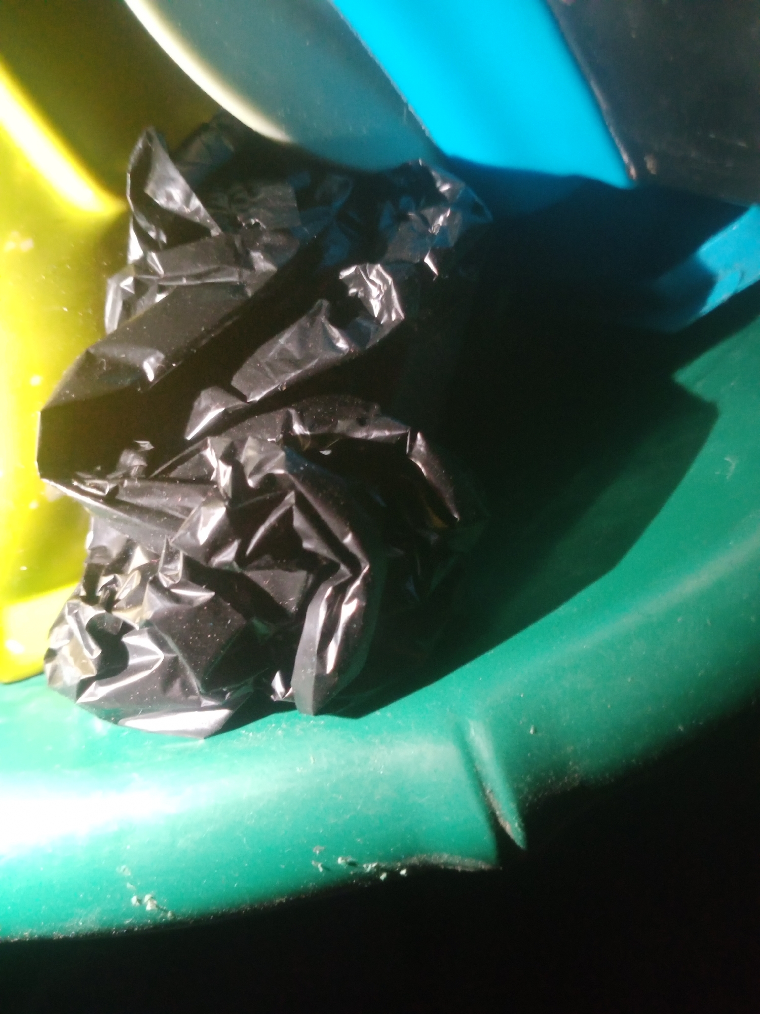 A crumpled black plastic bag is the central subject of this close-up image. The bag is situated on a green, curved surface, which appears to be part of a larger object, possibly a slide or a bin. To the left of the bag, a bright yellow surface is visible, and in the upper background, a portion of a bright blue surface can be seen. The lighting in the image creates strong shadows, particularly a large dark shadow cast by the bag and extending to the right. The green surface shows some dust or debris. The image lacks any indicators of its specific location within Jalingo, Nigeria, or any context regarding people or actions. The focus is entirely on the inanimate objects and their textures under dramatic lighting.