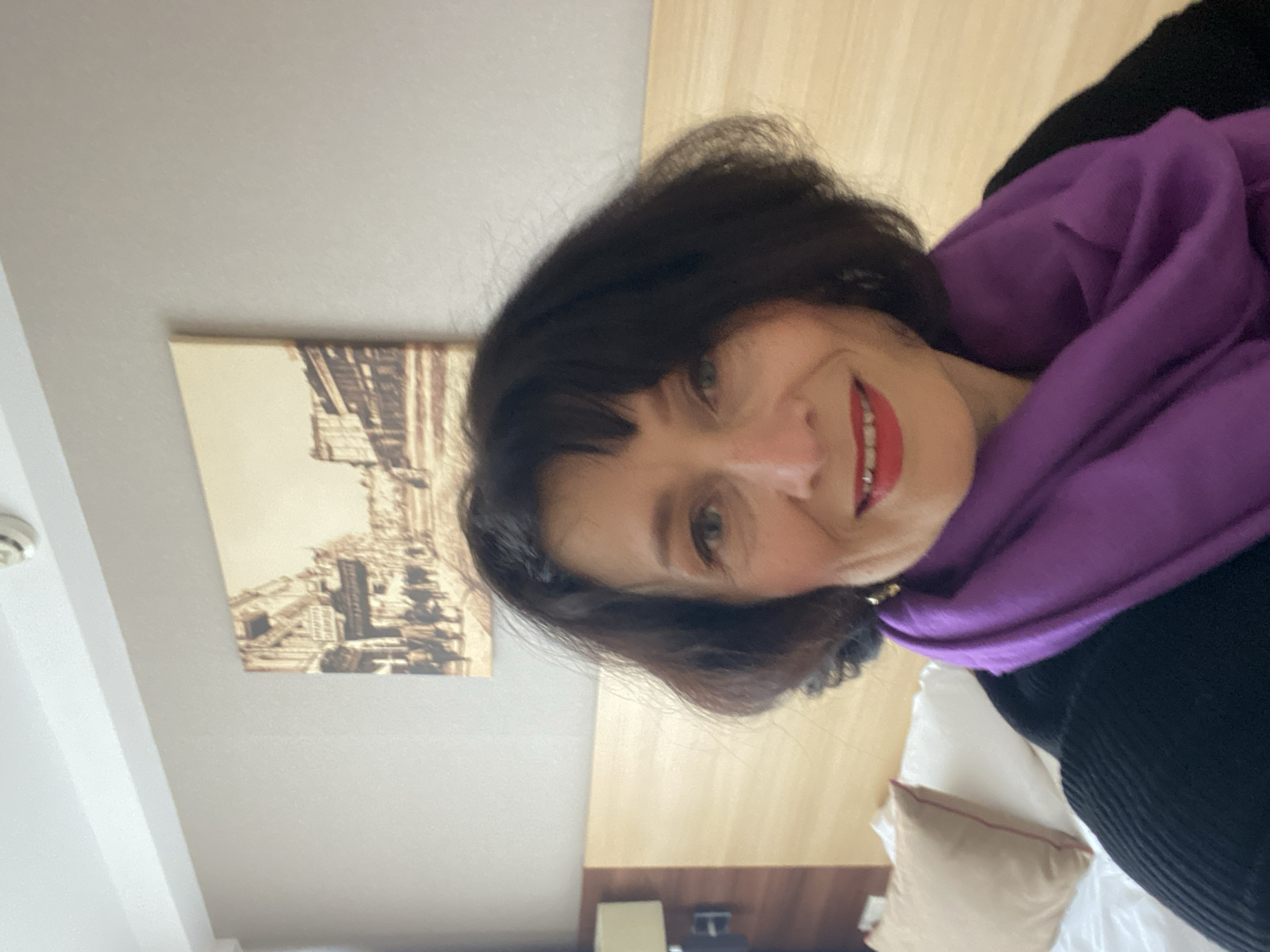 A woman with dark hair, red lipstick, and a purple scarf is smiling in a room. A framed sepia-toned image hangs on the wall, depicting an architectural scene reminiscent of ancient ruins, which suggests a location within a historic city; though it is impossible to verify the exact location of Kyiv based on the image content alone. The environment consists of a room with a bed, pillows, and a wood-paneled wall. There is no apparent interaction or specific action occurring other than the woman’s posed smile.