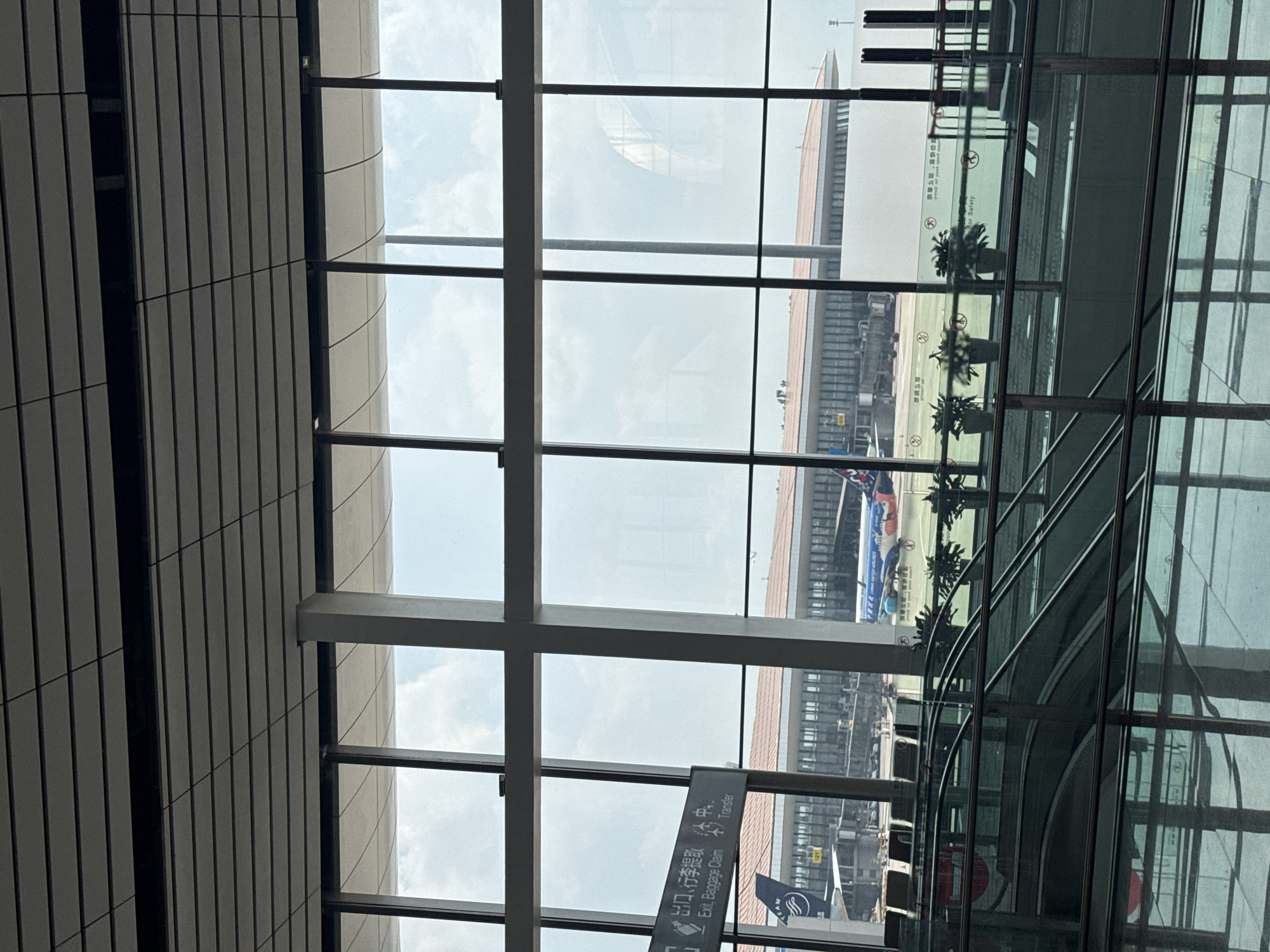 This image shows an interior view of a modern airport terminal, likely located in Guangyang District, China, judging by the signage. The perspective is from a high vantage point, looking out through a large glass window that offers a view of the tarmac and the sky. The window is divided by dark vertical and horizontal mullions, creating a grid-like pattern.
Inside the terminal, a section of a tiered, curved escalator is visible in the foreground, constructed with glass and metal railings. Above the escalator, a row of potted plants is arranged along a railing. Reflections of the interior, including more glass and the ceiling structure, are visible on the escalator's surfaces.
Outside the window, the airport tarmac is visible. An airplane with the branding "CHINA UNITED AIRLINES" and a colorful cartoon livery is parked. Another airplane, partially visible, has "TEAM" branding. Several jet bridges are connected to the terminal building, which has a distinctive curved roofline and numerous windows. The sky is overcast with scattered clouds, and the overall lighting suggests it is daytime. Signage in both Chinese and English, including directions for "Exit, Baggage Claim" and "Transfer," is visible on a sign mounted on a pole. The environment appears clean and well-maintained, characteristic of a contemporary airport.