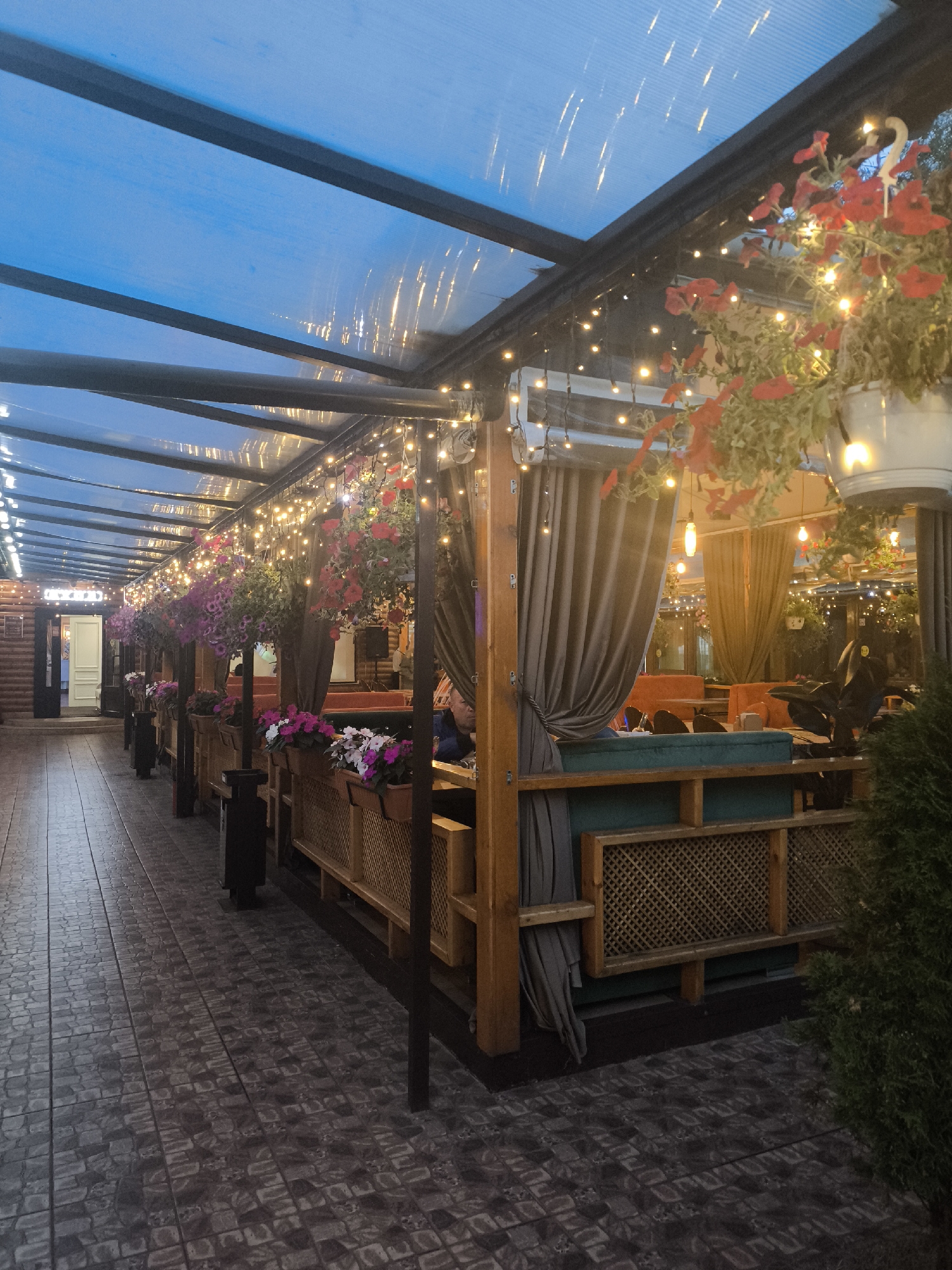 The image depicts an outdoor seating area of a restaurant or cafe in Khimki, Russia, during the evening. The setting is covered by a translucent, ribbed roof structure, illuminated by strings of warm-toned fairy lights that cast a cozy glow. Hanging flower baskets with red and pink petunias add vibrant splashes of color to the scene.
The seating area features wooden structures with ornate latticework, draped with neutral-colored curtains, creating semi-private booths. Orange and teal upholstered seating is visible within these booths. A tiled walkway runs along the length of the seating area.
In the background, more seating areas with similar lighting and floral decorations are visible, suggesting a larger establishment. A person is partially visible in one of the booths, engaged in a conversation or enjoying a meal. The time of day is twilight, indicated by the dim ambient light and the illuminated artificial lights. The weather appears to be clear and calm, suitable for outdoor dining.
A sign with Cyrillic text, possibly the name of the establishment, is partially visible on a wooden wall in the distance on the left side of the image. The overall mood is inviting and relaxing, with the lighting and floral arrangements contributing to a pleasant atmosphere.