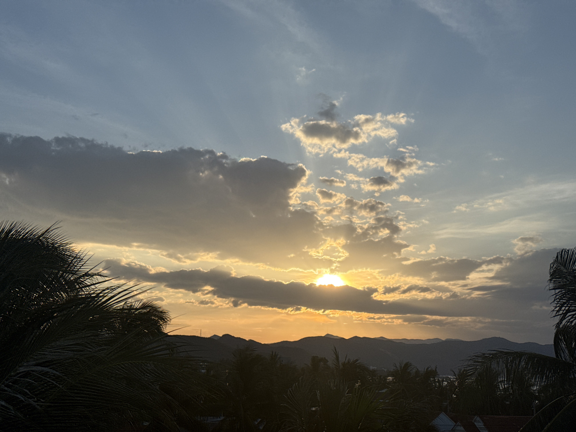 An outdoor, eye-level, medium shot captures a scenic sunset over a mountainous landscape. The sky transitions from a pale blue at the top to a warm orange hue near the horizon, illuminated by the setting sun peeking through a band of clouds. Rays of light pierce through the clouds, creating a dramatic effect. Silhouetted palm trees dominate the foreground, their fronds stretching across the lower portion of the frame. Beyond the trees, a range of dark, undulating mountains forms the middle ground, with a hint of a distant settlement visible at their base. The overall impression is one of natural beauty and tranquility, with the encroaching darkness of twilight.