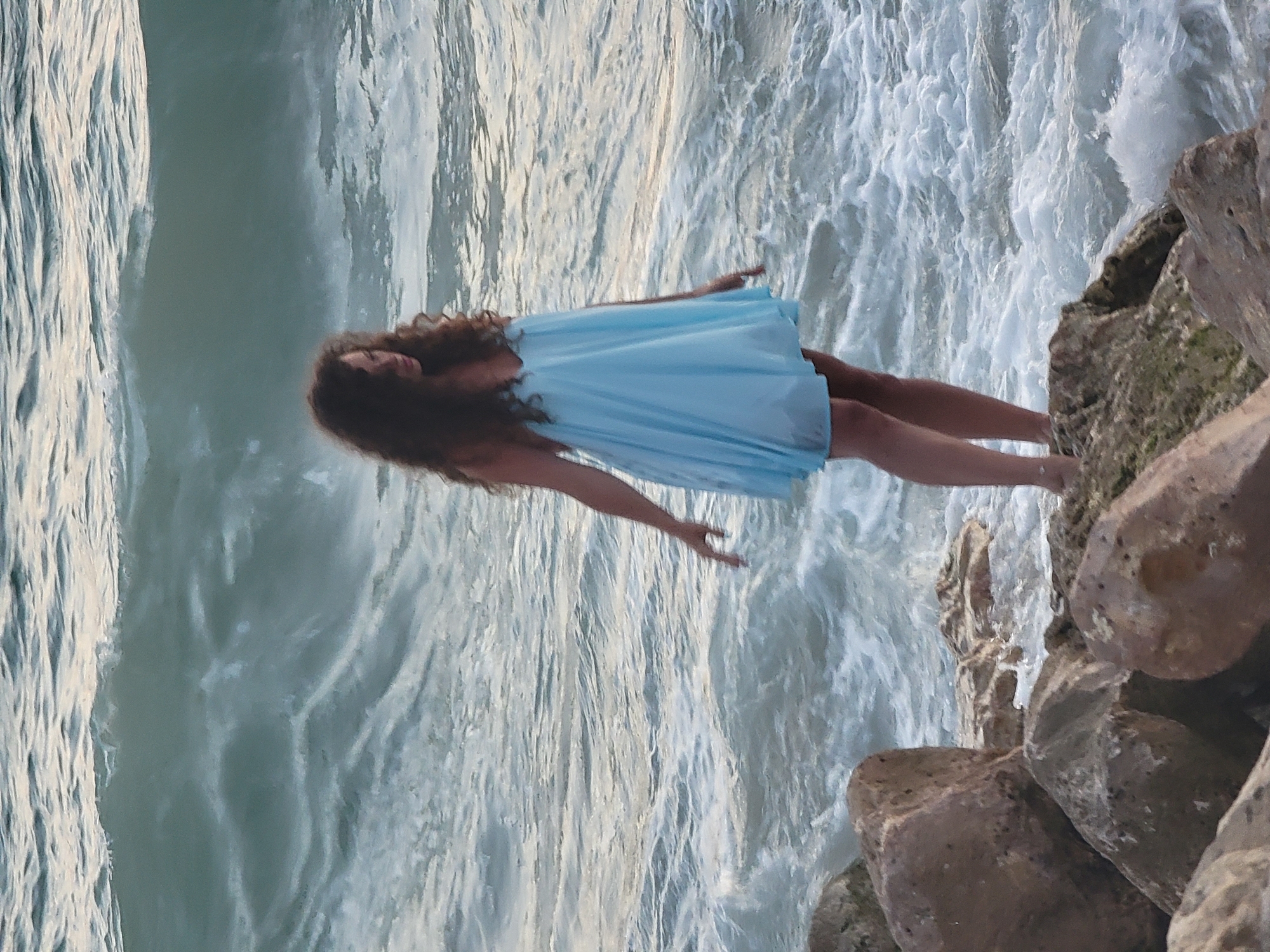 A woman with long, dark, curly hair, wearing a light blue, flowing dress, stands on large rocks at the edge of the ocean. She is facing towards the left of the frame, with her right arm extended downwards, her hand lightly touching the water. The ocean behind her is choppy with white waves crashing. The scene appears to be on a coastline, possibly in Quarteira, Portugal, given the rocky shore and the characteristic waves of the Atlantic Ocean. The lighting suggests it may be late afternoon or early evening.