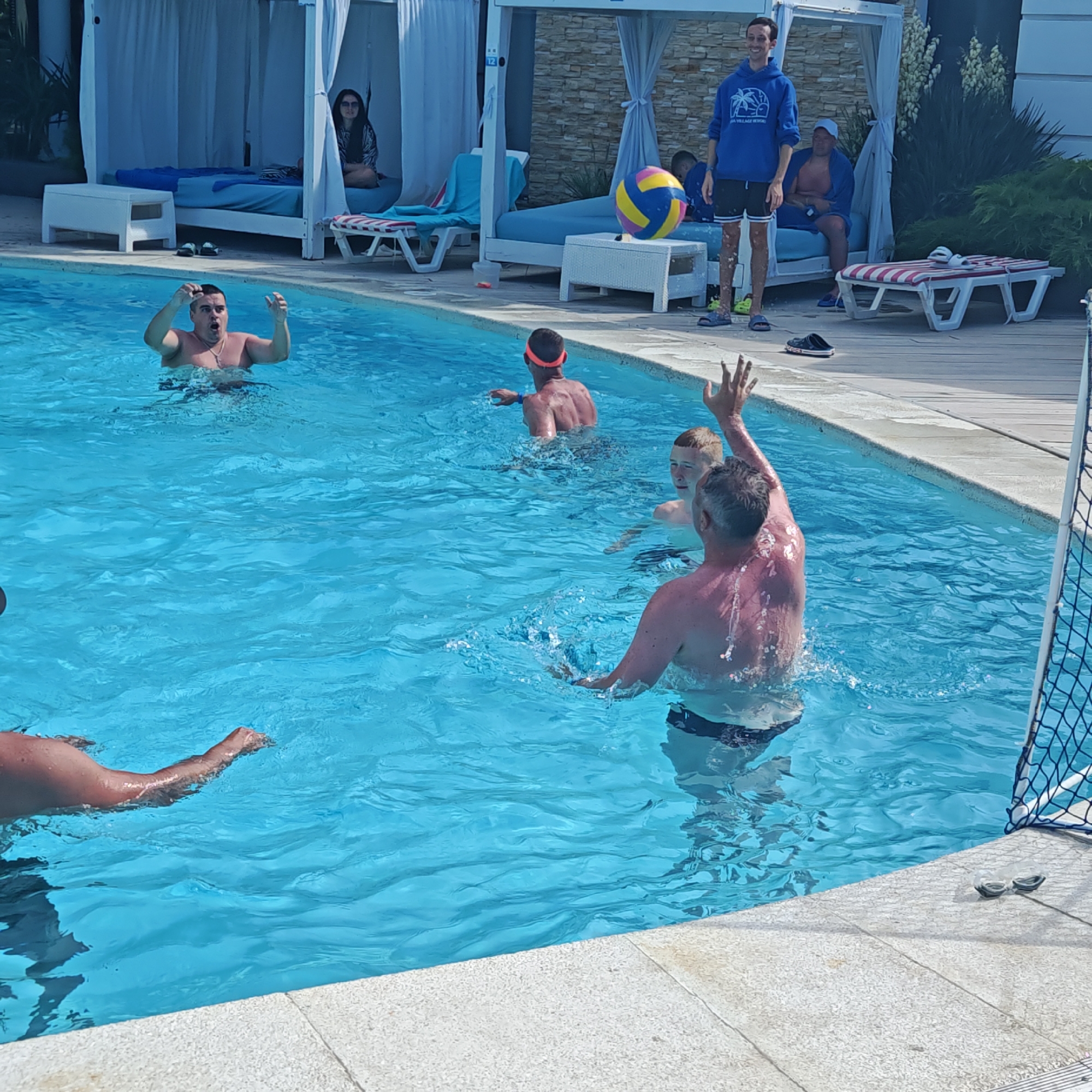 In a bright outdoor scene, a group of people are enjoying a game of water polo in a clear blue swimming pool. The sun is shining, indicating it's daytime, and the weather appears to be warm and pleasant. The pool is surrounded by a paved deck and lounge areas with white canopies, suggesting a resort or hotel setting.
Several individuals are actively participating in the game. One man in the foreground is reaching up with his hand, seemingly to block a shot or make a pass. Another man, slightly further back, is also in a throwing or reaching posture. Two younger individuals are also in the water, one with an orange headband, and another boy looking towards the action. In the background, a person stands by the pool wearing a blue hoodie and shorts, observing the game, while another person sits on a lounge chair. A woman is seated in one of the shaded cabanas in the background.
A colorful water polo ball is visible in the air, mid-game. Goggles and a water polo net are also present, further confirming the activity. The overall mood seems to be one of active recreation and enjoyment. The location is specified as Shcholkine, Ukraine.
