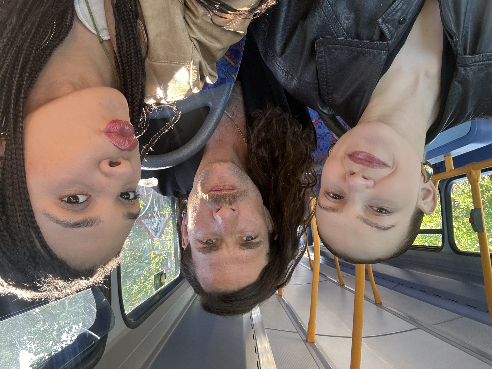 A group selfie features three individuals – two women and one man – positioned upside down within what appears to be a public bus. The subjects' faces fill the upper half of the frame, with their heads angled downwards towards the bottom of the image, suggesting they are leaning back or lying down while taking the photo.
On the left, a woman with dark braided hair and glossy, reddish-pink lips looks directly at the camera. In the center, a man with long, dark hair, a mustache, and a goatee stares intently. To the right, a second woman with a subtle smile and similar reddish lips also faces the camera. Both women wear dark clothing, with one in a beige jacket and the other in a black leather jacket. The man is also wearing a black leather jacket.
The setting is clearly the interior of a bus, characterized by light blue seats with a patterned fabric, distinct yellow/orange vertical poles, and large windows. Through the windows, bright sunlight illuminates green foliage, indicating a daytime setting, likely sunny. A triangular road sign, characteristic of UK warning signs (possibly indicating a double bend), is visible through the window behind the man. A steering wheel is partially visible near the man, suggesting they might be near the front of the bus. The overall atmosphere appears relaxed and informal. No discernible text is visible on the bus interior or on the subjects. Given the context provided, the location is London, United Kingdom.