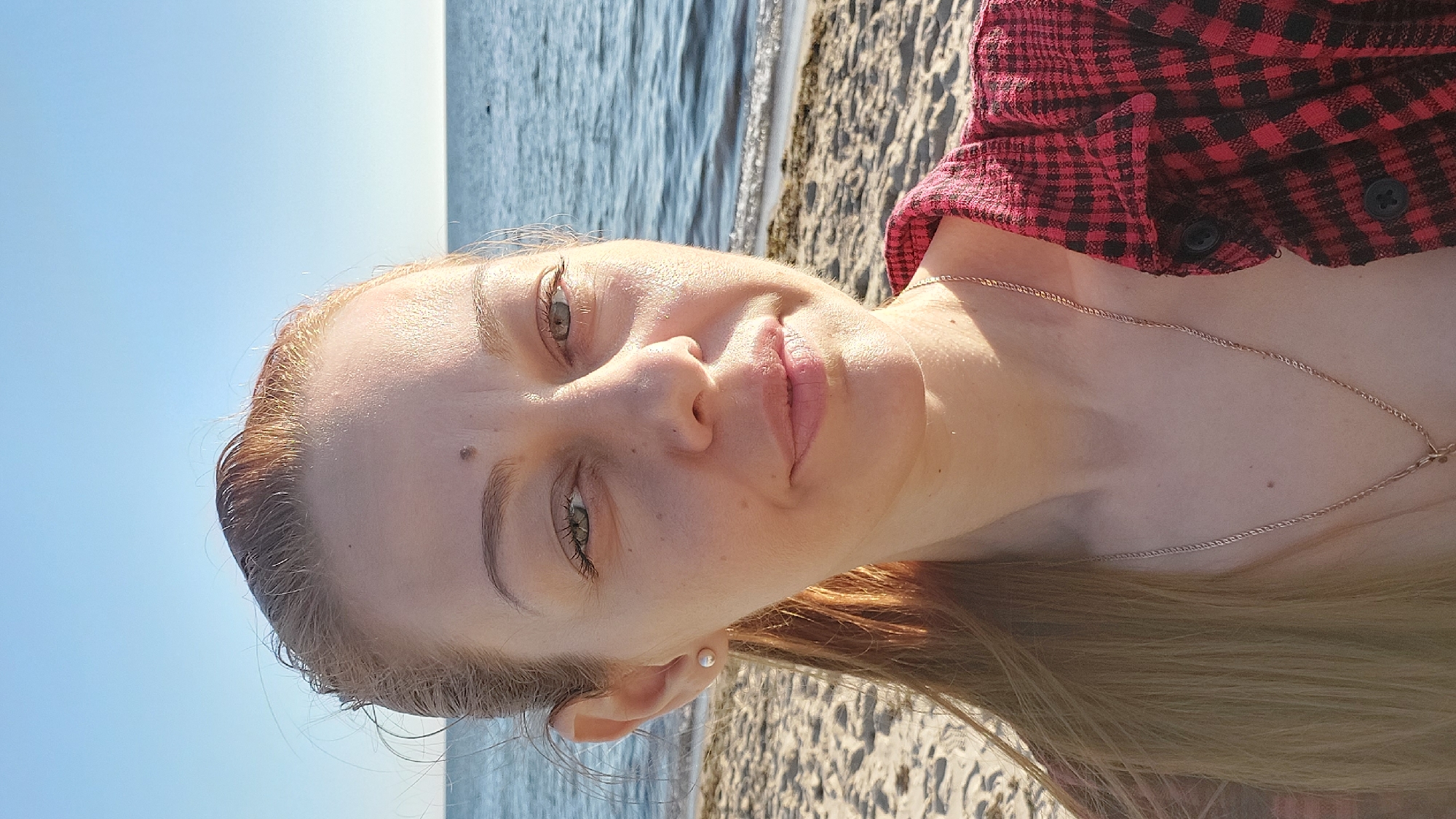 A blonde woman with fair skin and green eyes takes a selfie on a sandy beach, with the Baltic Sea and a clear blue sky in the background. She is wearing a red and black checkered shirt and a thin gold necklace. Her hair is pulled back into a ponytail. The sun is shining, casting shadows on her face. The location is Laulasmaa, Estonia.