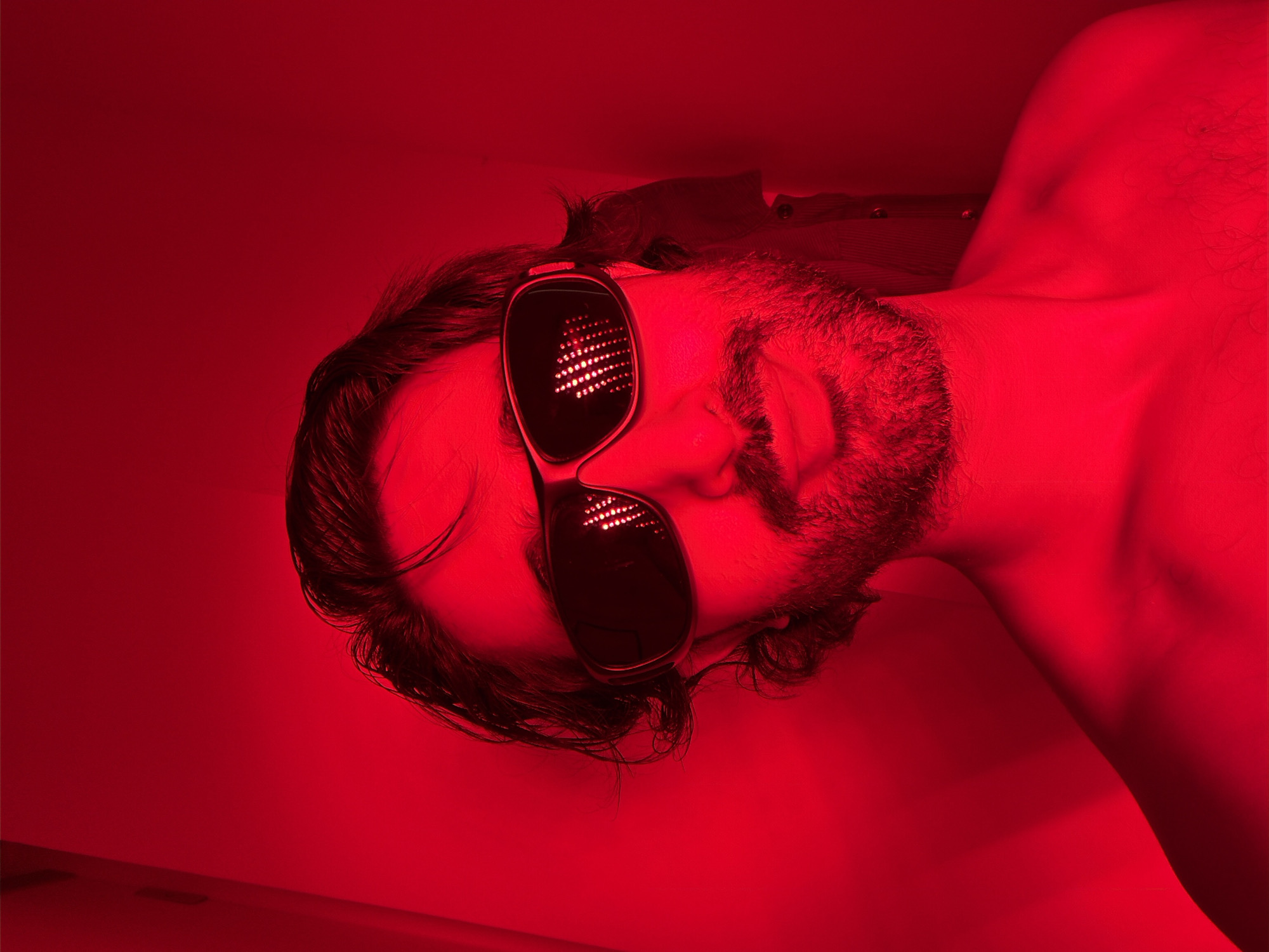 A man lies on his side, illuminated by red light. He is wearing sunglasses with a matrix of small lights reflecting off the lenses. He has dark hair and a beard. His shoulder and neck are visible. A portion of his shirt can be seen beneath his head. The scene is entirely bathed in a monochromatic red light. There is no information to indicate the location is in the city of London, United Kingdom.