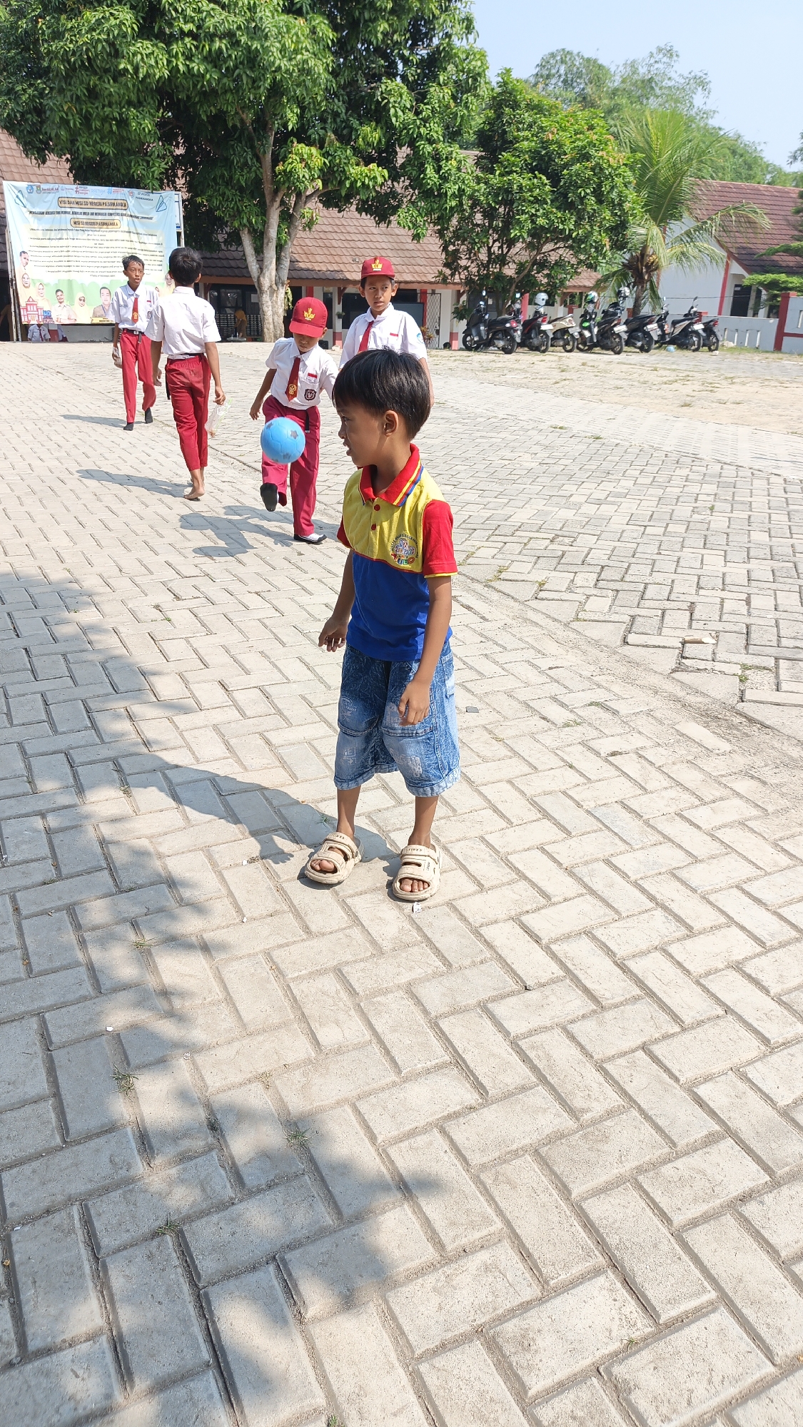 The image shows a group of children in a schoolyard in Pasir Bolang, Indonesia. The children are wearing school uniforms, consisting of white shirts, red pants, and red caps. One child in the foreground is wearing a blue and yellow shirt and denim shorts. Another child in the middle is kicking a blue ball. In the background, there are buildings, trees, and several motorcycles parked. The ground is paved with interlocking bricks. The scene appears to be a sunny day, with shadows cast by the children and trees. A banner with text and images is visible on the left side of the image.