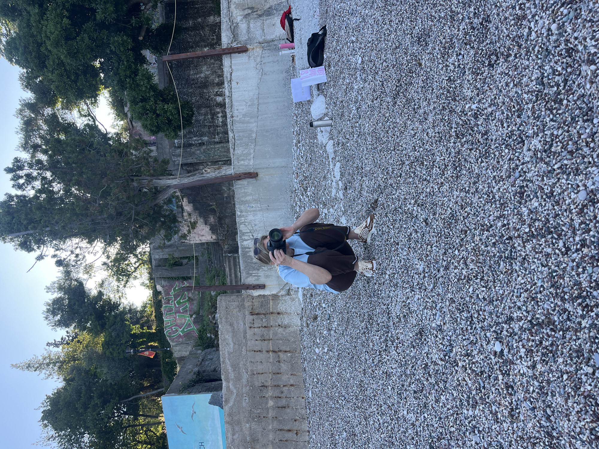 A person is squatting on a pebble beach with a camera in their hands, likely taking a picture. A concrete wall borders the beach. Behind the wall is a building with graffiti and some overgrown foliage. Further back there is a red hat, bag and a Dior shopping bag. The location is Boreti, Montenegro.