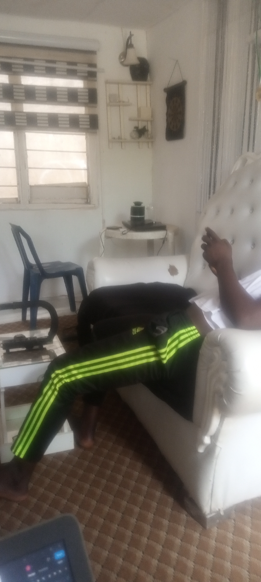 The image depicts a person seated in a white, tufted armchair within a room. The individual is wearing dark-colored Adidas track pants with three prominent neon green stripes down the side. They are partially obscured by a black jacket. A blue plastic chair is near a white side table, upon which rests a black object and a white surface. A window with horizontal blinds is visible in the background, along with a white wall adorned with a wall lamp, decorative shelves, and a small hanging tapestry. The floor is covered in a brown, diamond-patterned rug. Part of a tablet device is visible in the lower-left corner of the image.