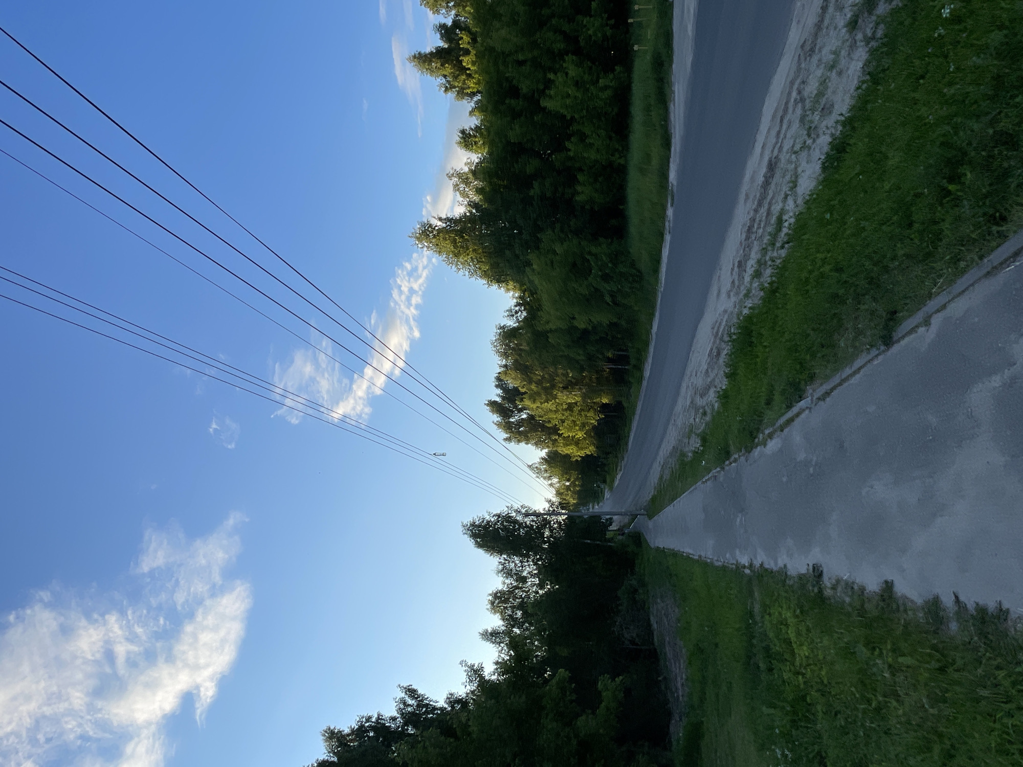 The image depicts a scene on a paved road in Krasnye Baki, Russia. The road curves to the left and ascends into the distance. On the left side of the road is a paved sidewalk, bordered by a strip of grass. Beyond the grass, a dense line of trees forms a dark green barrier. On the right side of the road, there is also a grassy verge, and beyond that, more trees, some of which are illuminated by sunlight. Overhead, several power lines stretch across the bright blue sky, which is dotted with scattered white clouds. The overall impression is one of a suburban or rural road, likely in the late afternoon or early evening, given the lighting. There are no people or vehicles visible in the image.