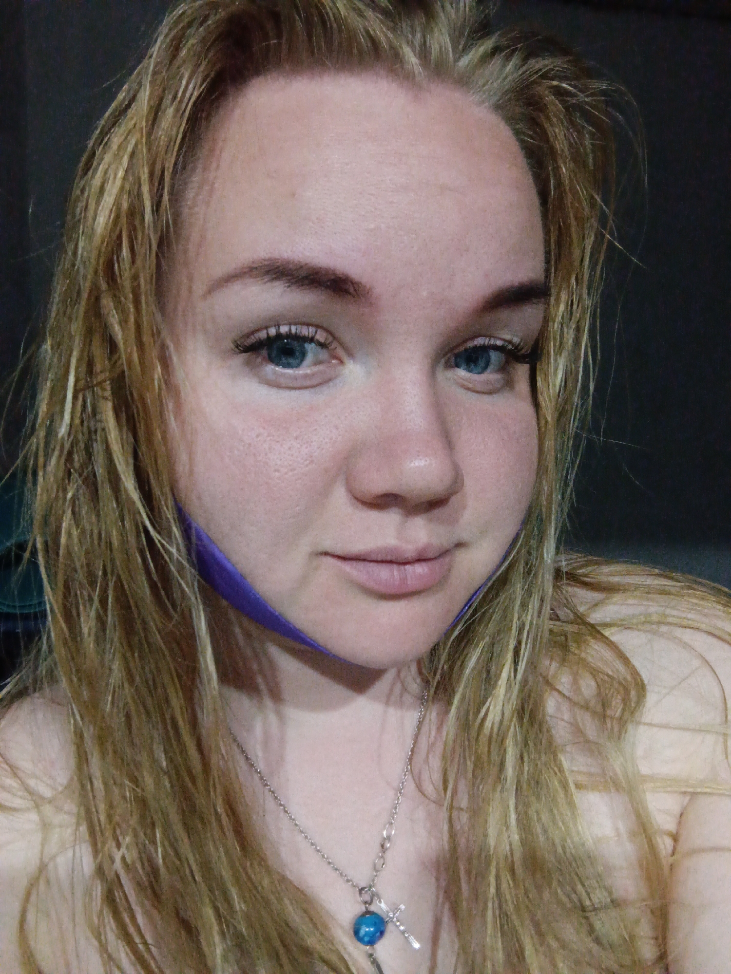 A close-up portrait of a fair-skinned woman with light blonde hair, likely indoors against a dark background. Her hair appears wet or damp and is framing her face. She has blue eyes and light eyebrows, and is wearing subtle eye makeup including mascara or false eyelashes. A thin, purple fabric or ribbon is visible draped down one side of her face, possibly indicating it was once attached to a face mask. She is wearing a silver necklace with a cross pendant, and a blue gemstone at the base of the cross. She is not showing any specific city of Фоки, Russia landmarks.