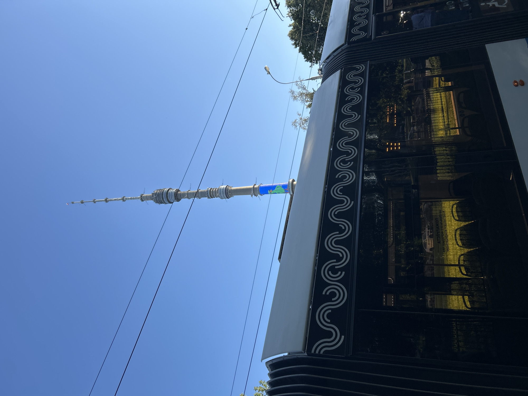 The image displays a close-up, low-angle view of a television tower against a clear blue sky. The tower appears to be the Ostankino Tower in Moscow, Russia. The lower section of the tower, visible in the frame, has a distinctive segmented design with lights. A blue section with what looks like a map or graphic is visible partway up the tower. In the foreground, the side of a building or vehicle with dark, reflective glass and a decorative wavy pattern along its edge is prominent. The reflections in the glass suggest an outdoor setting, possibly a park or a street with trees and sunlight. The time of day appears to be daytime, indicated by the bright blue sky and the sunlight casting reflections. No people are clearly visible, but the reflections in the building suggest some presence inside or nearby. The overall scene conveys a bright, clear day in an urban or semi-urban environment. Visible text includes a partial sign in the reflection of the building that appears to be in Cyrillic script.