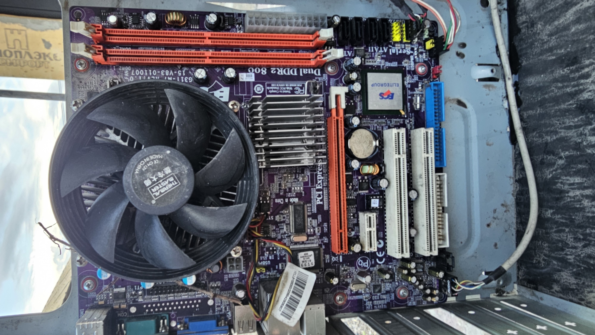 This image shows a close-up, overhead view of an open computer motherboard with its CPU cooler fan prominently displayed. The motherboard, which is purple with various components, appears to be situated within a computer case, suggesting it's part of a desktop computer. The scene is outdoors, indicated by the bright sky and what looks like a blurry background of clouds and possibly structures. The lighting suggests it might be daytime. There are no people visible in the image.
The motherboard itself is the primary subject, with visible text on it including "Dual DDR2 800," "PCI Express," "ELITEGROUP," and "THERMAL MASTER." There is also Chinese text which translates to "Cool Master." The motherboard has several slots for RAM, expansion cards, and various chips and capacitors. The CPU cooler fan is large and black, with "THERMAL MASTER" printed on its center. Cables are connected to some components.
The overall impression is that of a disassembled computer component, possibly being examined or prepared for repair or recycling, given the outdoor setting and the open nature of the computer case. The setting appears somewhat industrial or utilitarian, with a rough, textured surface visible in the background to the right and a metallic, possibly weathered, structure to the left. No specific event is depicted, but the image focuses on the technical details of the computer hardware.