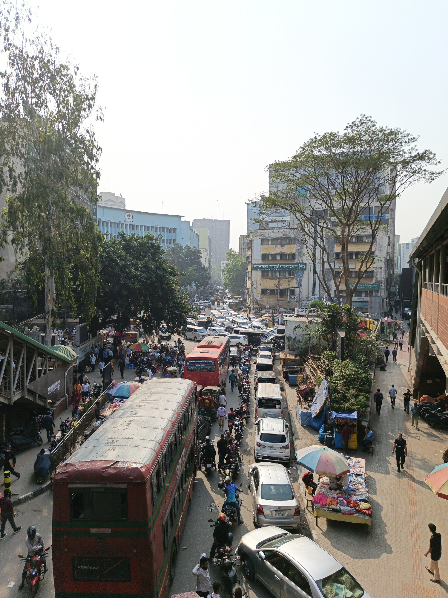 The image depicts a bustling street scene in Dhaka, Bangladesh, likely near the Motijheel area. The view is captured from an elevated perspective, possibly a bridge or high building. The street is crowded with various vehicles, including cars, motorcycles, and buses, indicating heavy traffic. The sidewalks are filled with pedestrians, many of whom are walking alongside street vendors selling goods under colorful umbrellas. Several buildings surround the street.