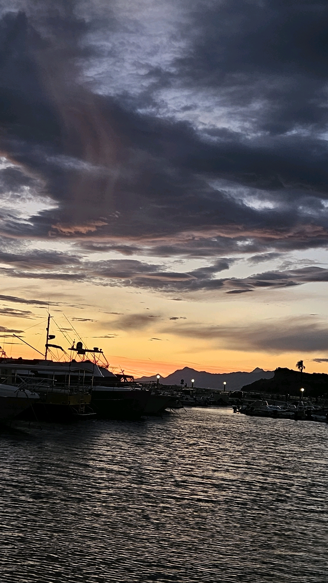 The image depicts a harbor scene at dusk in Općina Tučepi, Croatia. The sky is filled with dramatic, dark clouds tinged with hues of pink and orange from the setting sun. In the foreground, the dark water of the harbor is rippled, reflecting the twilight sky. Several boats are docked, with their silhouettes prominent against the colorful sky. The boats vary in size, with at least one larger vessel featuring a mast and rigging. In the background, a silhouetted mountain range is visible, with a single palm tree standing on a dark hillside to the right. Small, illuminated lamps are dotted along the waterfront. The overall atmosphere is serene and tranquil, characteristic of a quiet evening in a coastal town. There are no visible people or animals in the image.