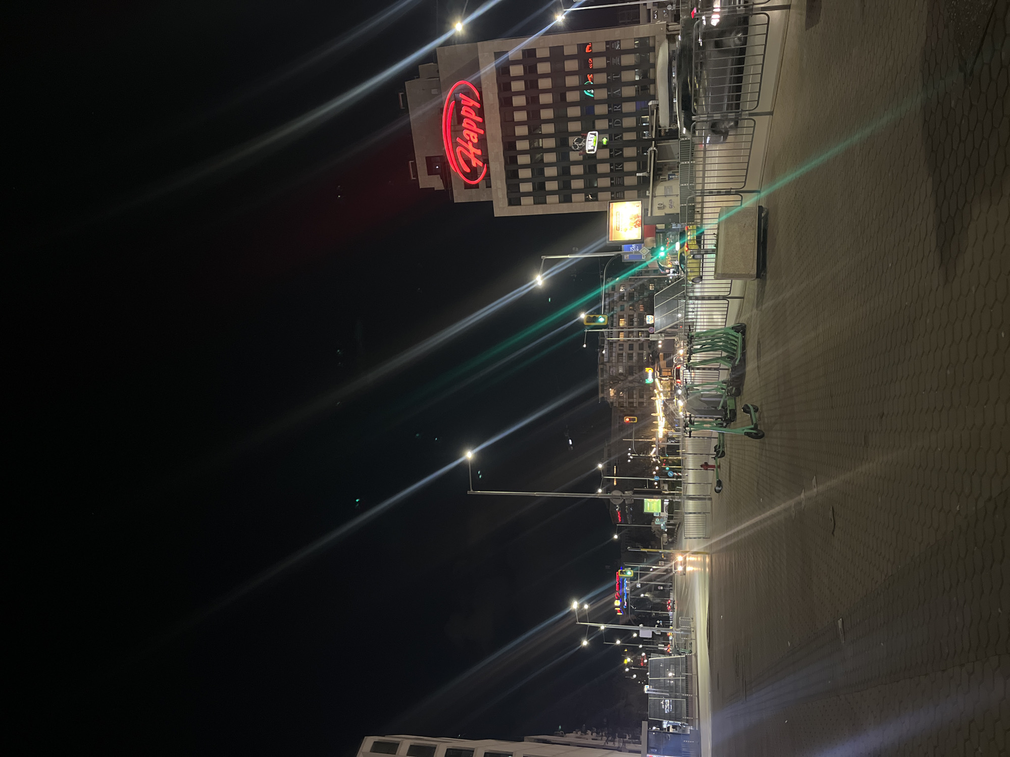 The image depicts a street scene at night in Sofia, Bulgaria. The view is along a paved walkway lined with streetlights. On the right side of the walkway are several electric scooters parked in a rack. In the background, a building prominently displays a large, illuminated "Happy" sign in red. Streetlights and traffic lights illuminate the street and surrounding buildings. The overall environment is urban with a mix of modern and older architecture. There are railings next to the sidewalk. There are no people visible in the picture.
