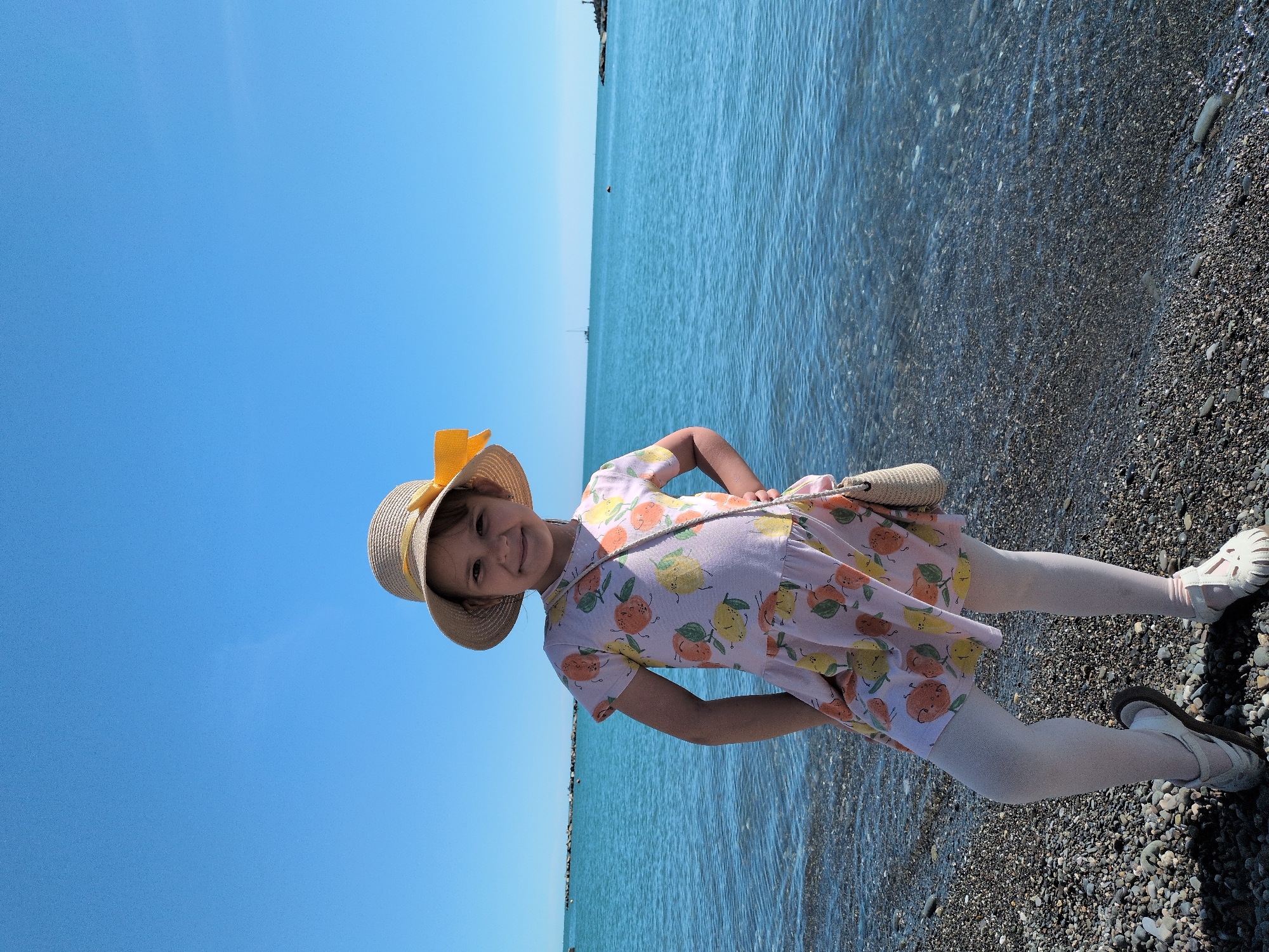 A young girl is standing on a pebble beach next to a large body of water, likely the Black Sea in Sochi, Russia. She is smiling cheerfully at the camera with her hands on her hips.
The girl is wearing a light-colored, short-sleeved dress printed with a pattern of cartoon-like oranges and lemons. On her head, she has a straw hat adorned with a bright yellow ribbon tied in a bow. She also wears light-colored tights or leggings and light-colored sandals. A small, woven, light-brown bag hangs over her left shoulder.
The scene depicts a clear and sunny day, likely midday or early afternoon, under a vast, bright blue sky. The horizon line is visible, separating the sky from the calm, blue-green water, which shows gentle ripples. The shore consists of dark grey pebbles, visible beneath her feet and extending along the water's edge. Faint outlines of distant land or structures can be seen along the horizon. The overall impression is one of a happy and pleasant day outdoors by the sea. There is no visible text in the image.