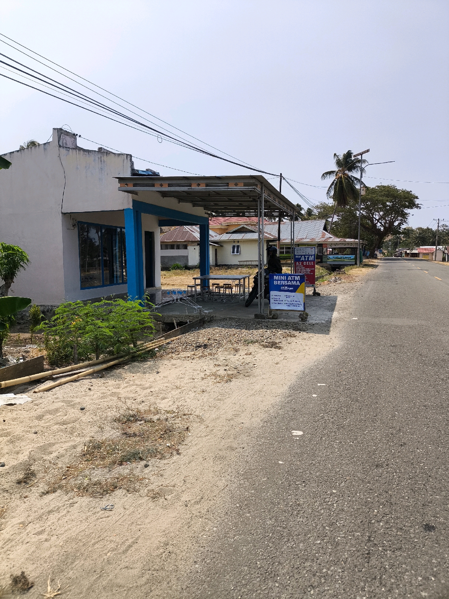 A white building with blue trim and a covered outdoor area is situated next to a paved road in Kwandang, Indonesia. The building appears to be a small business, possibly a convenience store or a service provider, with signs indicating "MINI ATM" and "AZ CELL". A motorcycle is parked under the covered area, and a person is seated on it. Several tables and chairs are set up outside the building, suggesting an outdoor seating area. The surroundings are dusty and dry, with some vegetation and bamboo poles visible. The sky is clear and bright, indicating a sunny day. The road stretches into the distance, with other buildings and trees lining its sides.