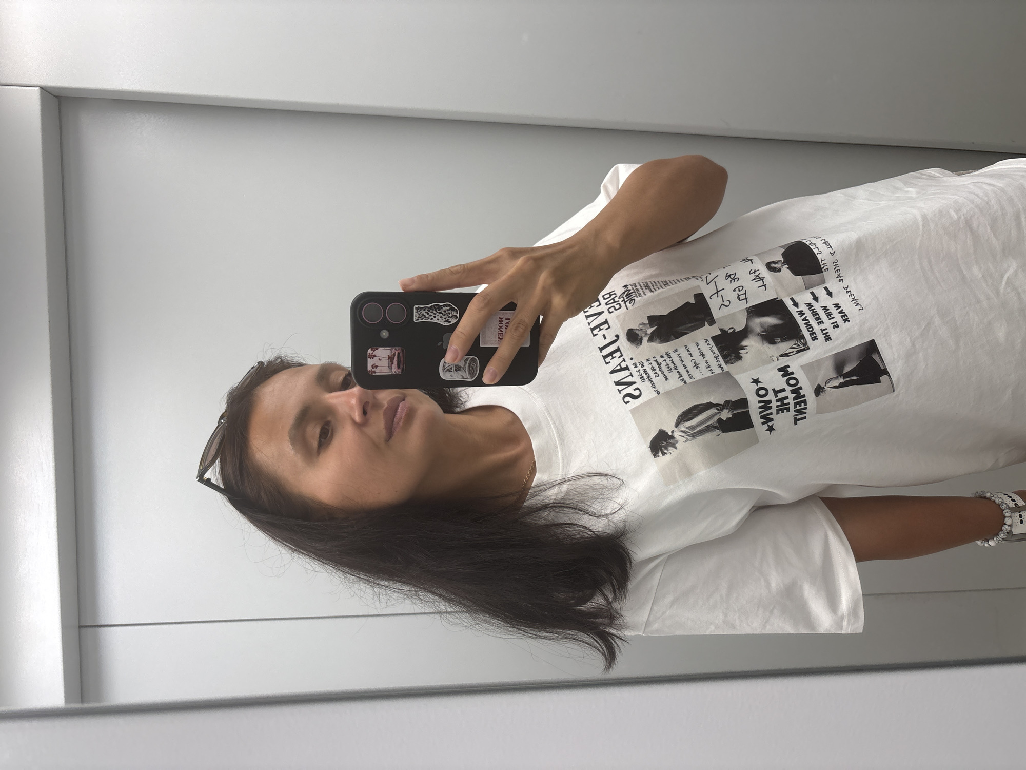 In a full-body mirror selfie, a woman stands in what appears to be an elevator, judging by the metallic doors and vertical lines of the structure. She has dark brown hair that falls past her shoulders and is wearing a white t-shirt with a collage of black and white images and text on the front. She is holding a black smartphone in her right hand, which is decorated with several stickers, including one of a pink vintage car, a leopard print pattern, a US dollar bill, and a grey and white abstract design. She also wears a silver bracelet with white beads on her left wrist. The background is a plain grey wall, suggesting an indoor setting. The woman's expression is neutral as she looks into the mirror. The image is well-lit, with no discernible shadows that would indicate the time of day or light source outside of the assumed indoor lighting. The location is identified as Chaikovsky, Russia, though there are no specific visual cues within the image to confirm this.