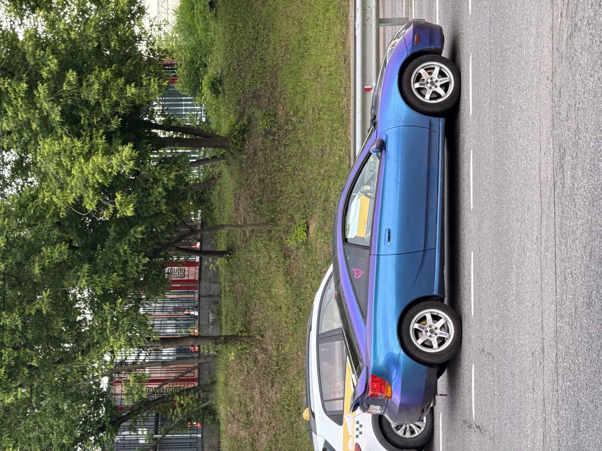 A blue sports car with a pink heart sticker on the rear window is parked on a road in Moscow, Russia. The car is in the foreground, and a white taxi with a yellow roof sign is visible behind it. The road is paved, and there are lane markings on the ground. In the background, there are trees and a fence. The overall impression is that of a typical urban street scene.