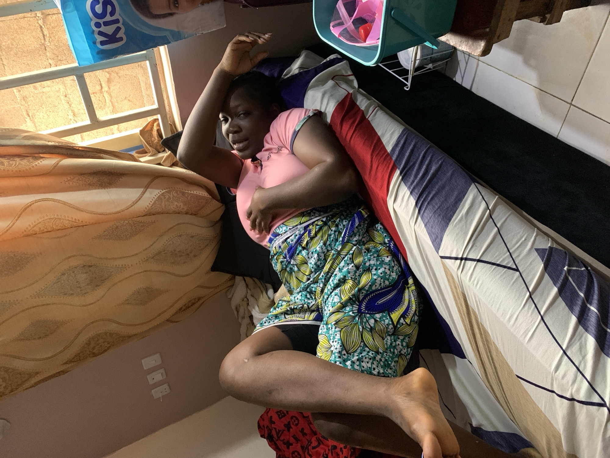 A person is lying on a bed in a room. The person is dark-skinned, wearing a pink shirt and a brightly patterned skirt. They are lying on their back with one arm raised above their head and the other across their chest. The bed has patterned sheets and a blanket with red, white, and blue stripes. A small, teal-colored plastic container is visible at the head of the bed. The room appears to have cream-colored walls and dark flooring. The location is Rafin Bauna, Nigeria.