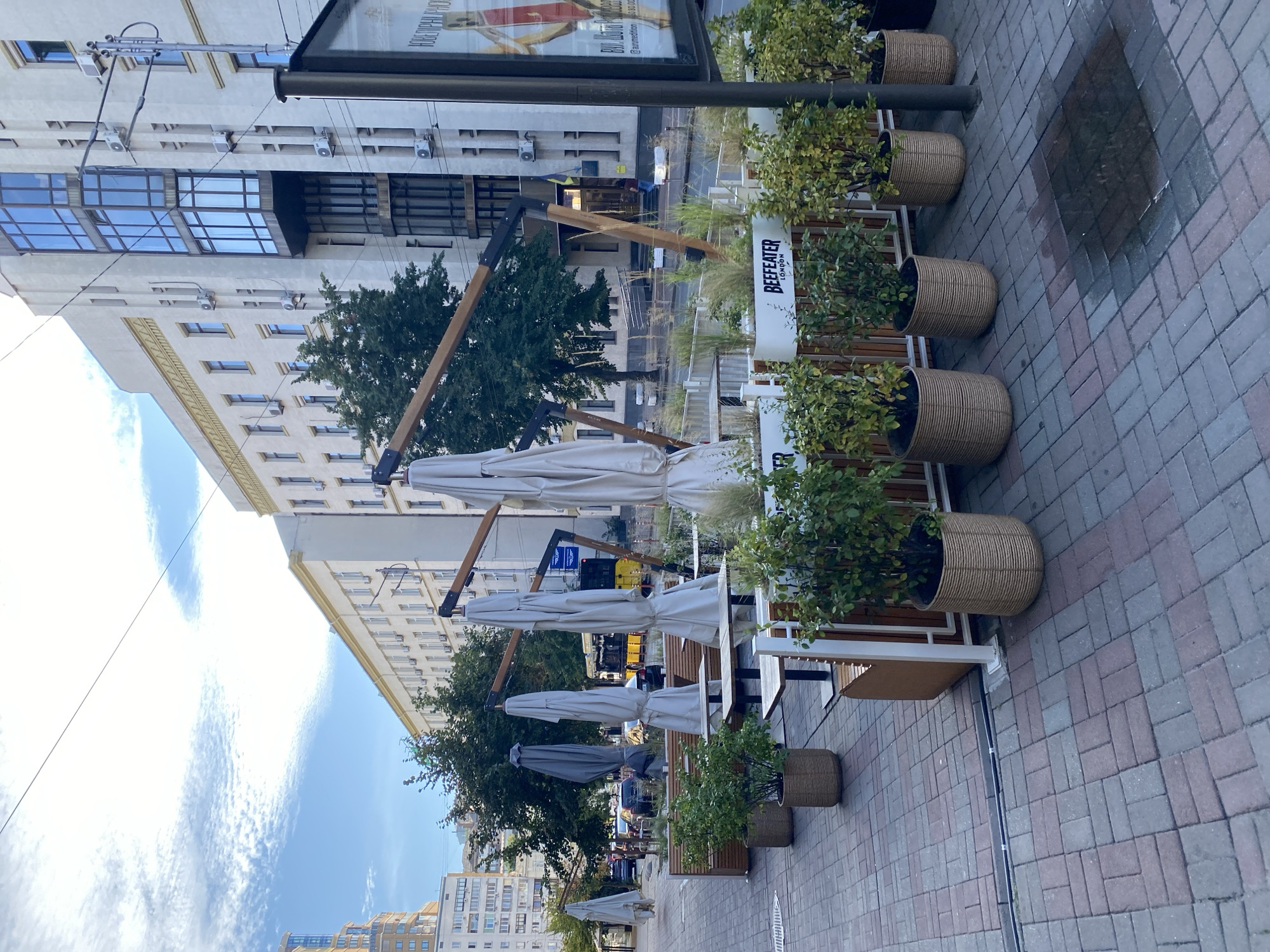 The image depicts an outdoor seating area of a restaurant or cafe on a city sidewalk. In the foreground, there are several tables and chairs, with closed umbrellas overhead supported by wooden structures. Potted plants in woven baskets line the perimeter of the seating area. A sign for "BEEFEATER LONDON" is visible on one of the planters. The sidewalk is paved with interlocking brick pavers. In the background, there are buildings, trees, parked cars, and a yellow tram. The sky is partly cloudy. The scene appears to be an urban environment.