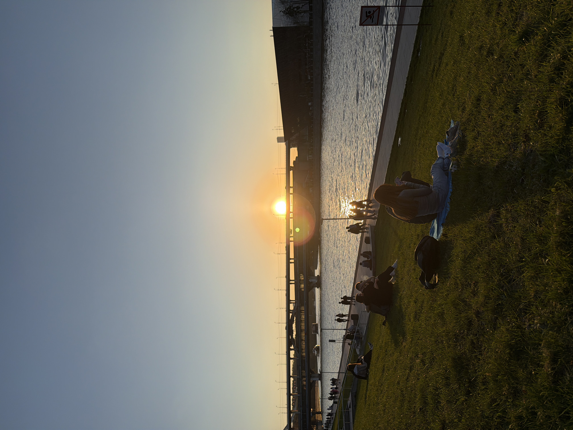 The image captures a sunset scene along a riverbank in Moscow, Russia. The sun is setting directly in line with a bridge over the river, creating a strong glare and lens flare in the image. Silhouetted against the setting sun is a bridge structure, possibly a railway bridge, with multiple supports and overhead lines. Along the river's edge, a concrete embankment transitions to a grassy area where several people are relaxing and enjoying the sunset. There are a few people lying on blankets, some sitting and others walking along the embankment. A sign post is visible near the grass. The sky is a gradient of blue to orange. The water reflects the sunlight, creating a shimmering effect. Overall, the scene depicts a peaceful evening atmosphere by the Moscow river.