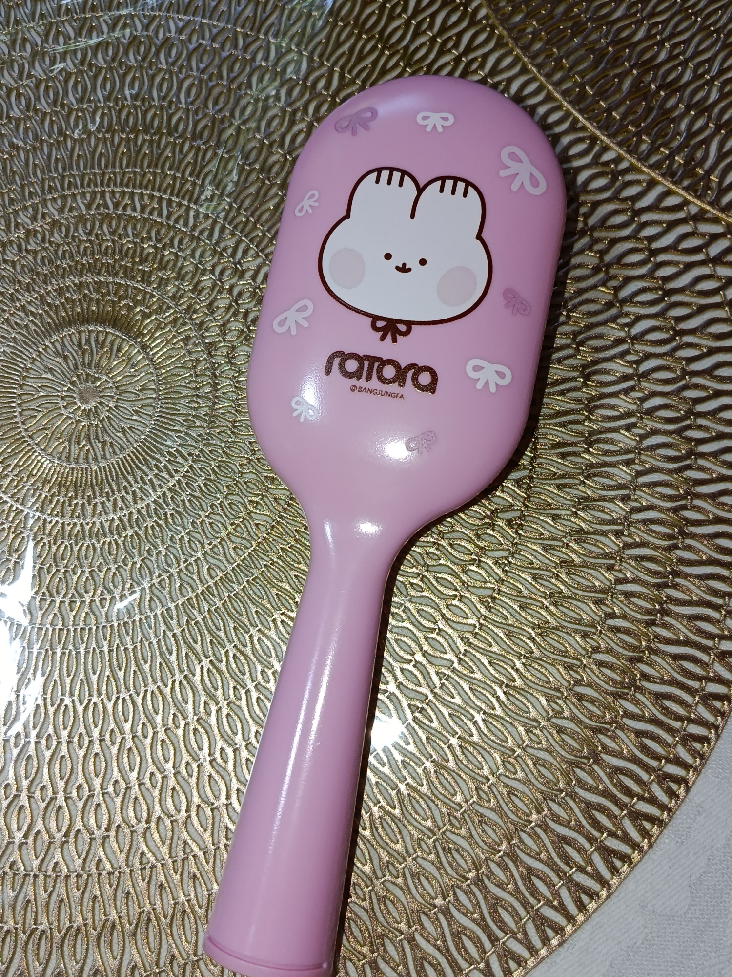 A pink hairbrush with a cartoon rabbit face and small bow accents rests on a textured gold placemat. The placemat has a circular, woven pattern. The hairbrush has the word "rara" and "@ SANGJUNGFIA" printed on it in a dark font. The image is a close-up shot, with the handle of the hairbrush extending downwards and out of frame. The background is the gold placemat. There are no people or indications of location in Tashkent, Uzbekistan.