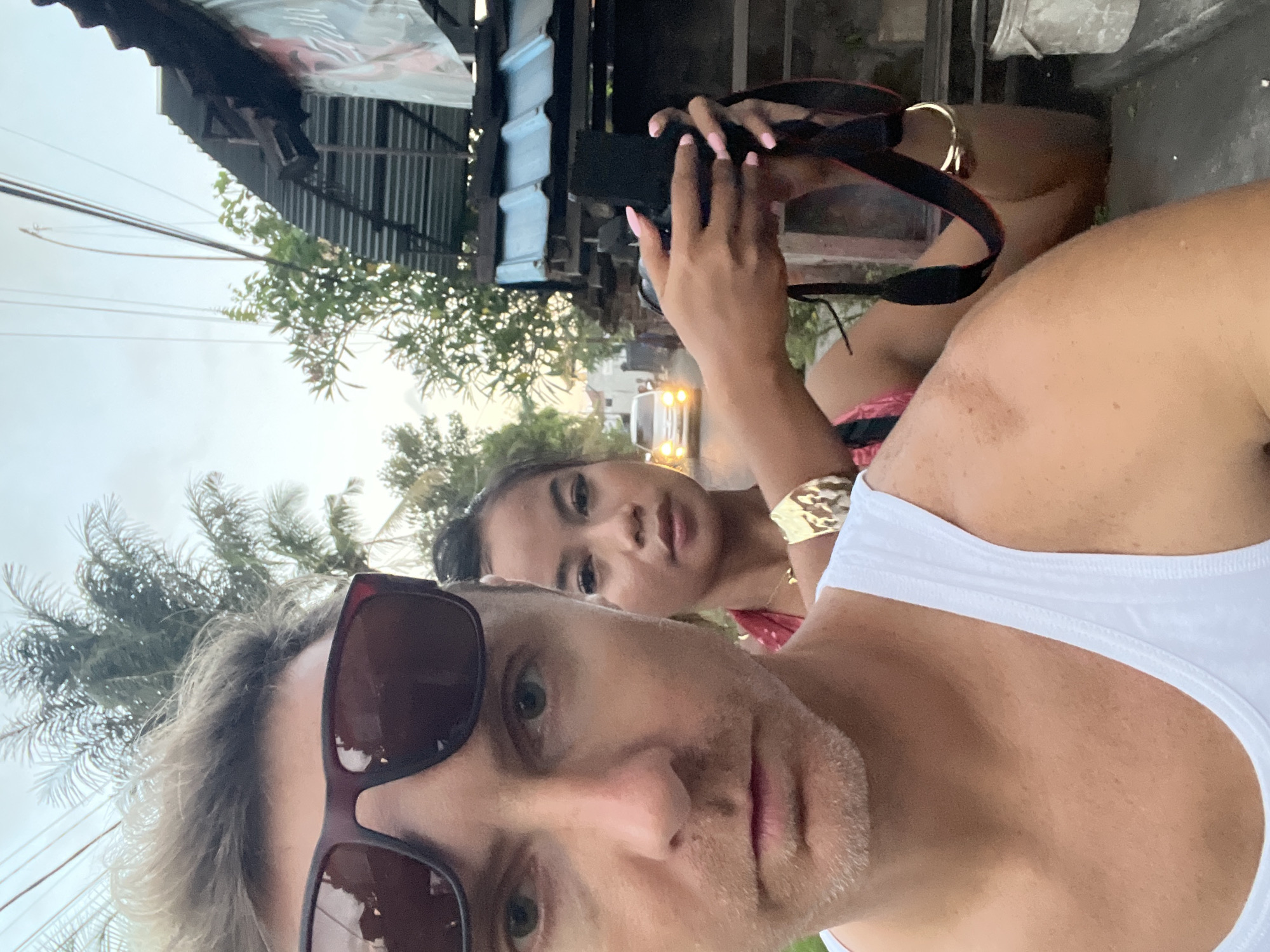A selfie shows two individuals on a street in Sukawati, Indonesia. The person in the foreground wears sunglasses and a white tank top. A woman stands behind him, holding a black camera with a strap, and wears a gold cuff bracelet. They stand on a paved area adjacent to a building with a corrugated roof. Green foliage is visible in the background, along with a street scene including a vehicle with headlights. The sky appears overcast.