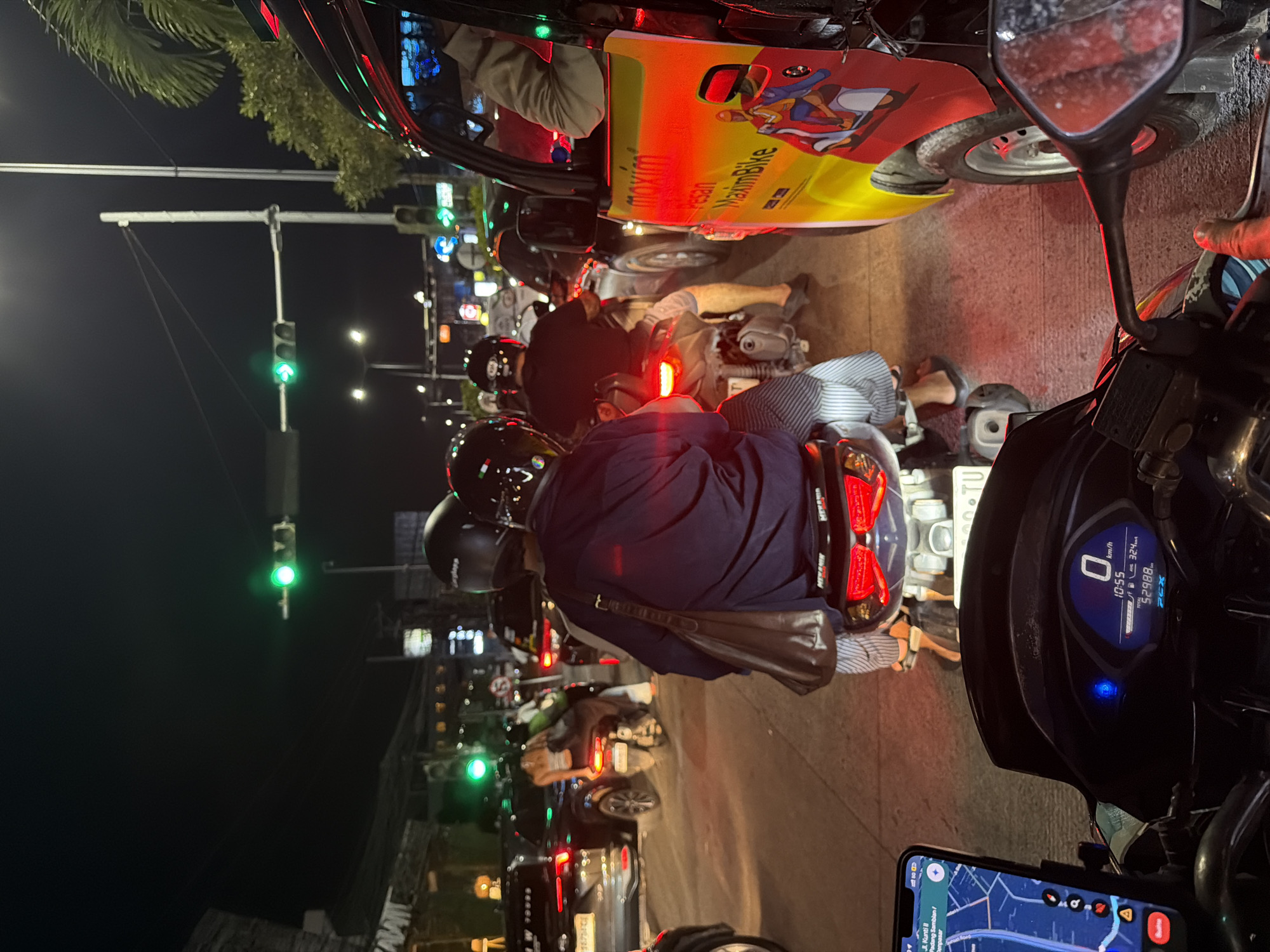 The image depicts a night scene in Kuta, Indonesia, with traffic congestion. A motorcycle rider, positioned behind other vehicles and motorcycles, is waiting at a traffic light. The traffic lights overhead show a green signal, but the vehicles are not moving. A Maxim Bike taxi is visible, as is a smartphone displaying navigation on the rider's motorcycle. The dashboard of the motorcycle is visible with the speedometer at zero, and the time 10:55. The road appears crowded, and street lights provide illumination.