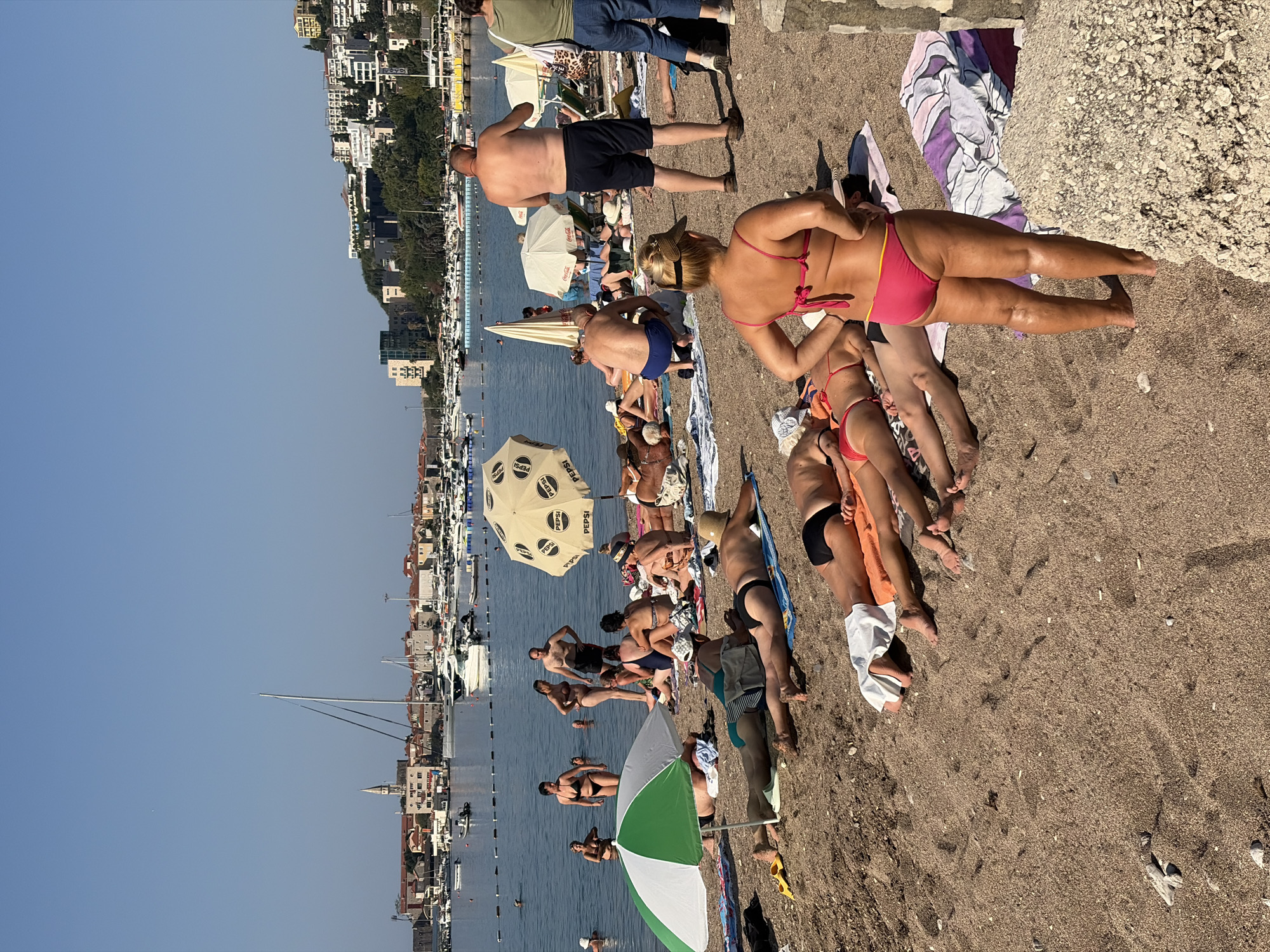 The image depicts a sunny day at a crowded beach in Budva, Montenegro, with the city's skyline and harbor visible in the background. Numerous people are sunbathing on the sand, some lying on towels and others in lounge chairs. Several individuals are in the water, while others are walking along the shoreline. Beach umbrellas, some with prominent branding, are scattered across the sand. The water is calm and blue, and several boats are docked in the harbor. The architecture in the background suggests a coastal Mediterranean town with red-tiled roofs and a church steeple. The overall scene conveys a typical summer vacation atmosphere.