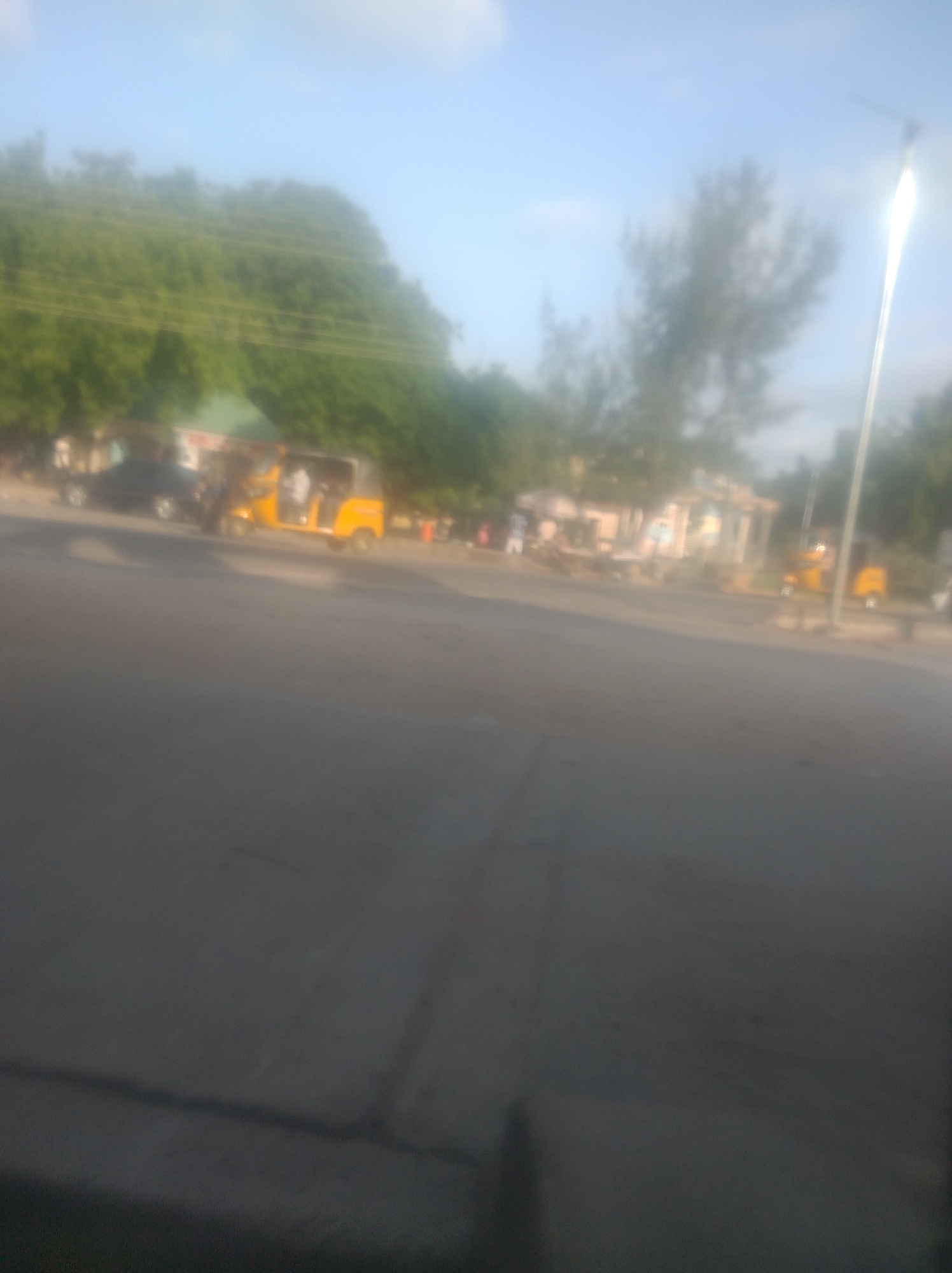 The image shows a street scene in Jalingo, Nigeria, with a clear sky and some trees in the background. In the middle ground, there are several vehicles, including a yellow auto-rickshaw and a dark-colored car. There are also some people visible, though their features are indistinct due to the image's blurriness. The foreground is dominated by a paved road. The scene appears to be a typical street environment in a town or city.