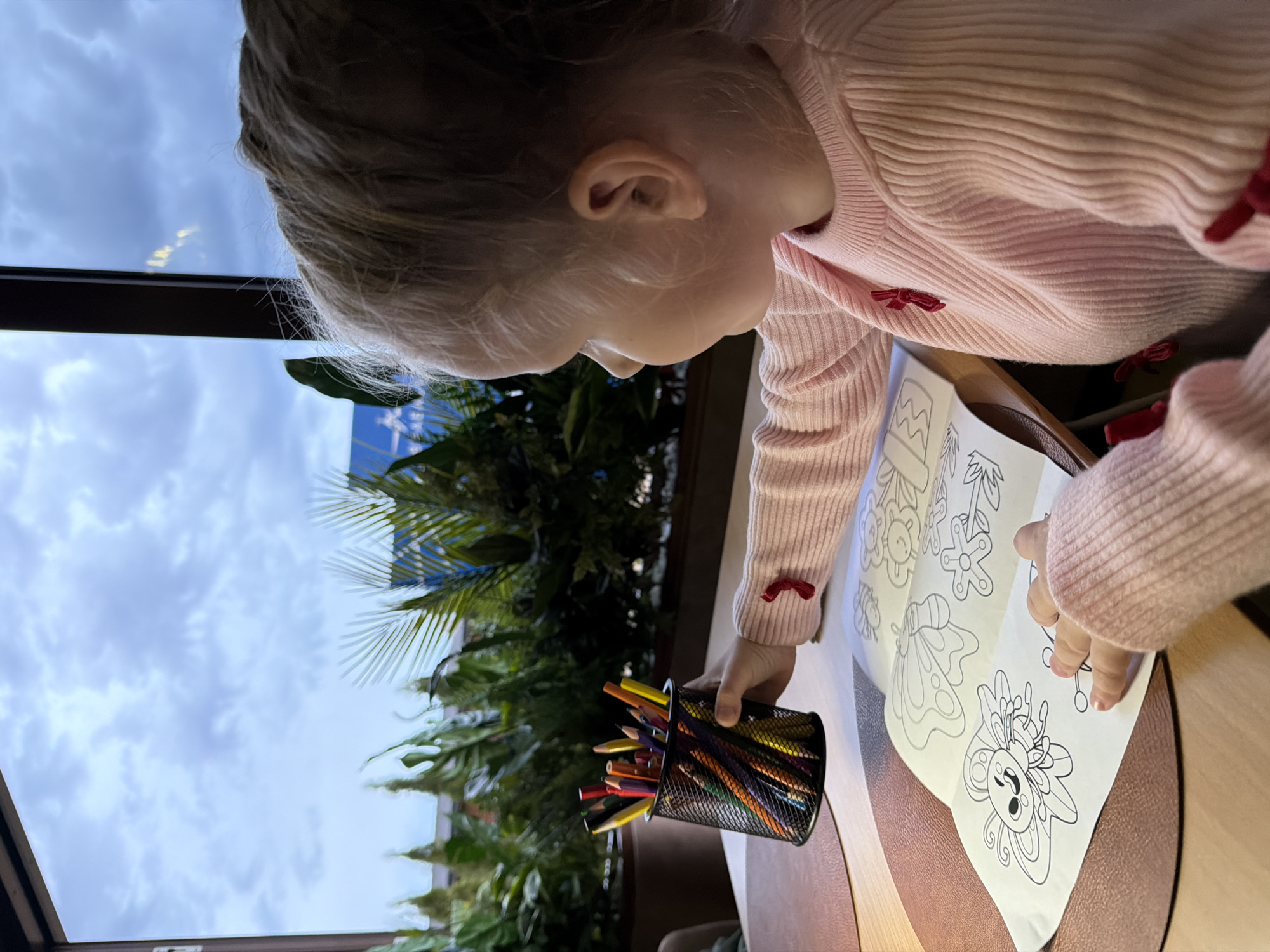 A young child with fair hair is seated at a round table with a coloring book open in front of them. The child is wearing a pink sweater with red bow accents. The table is made of light-colored wood with a brown, leather-like circular inlay. A black wire mesh container filled with colored pencils is positioned next to the coloring book, and the child's hand is extended towards the coloring book. Outside a window, there is a view of palm trees and a cloudy sky. The location is Kamyshin, Russia.