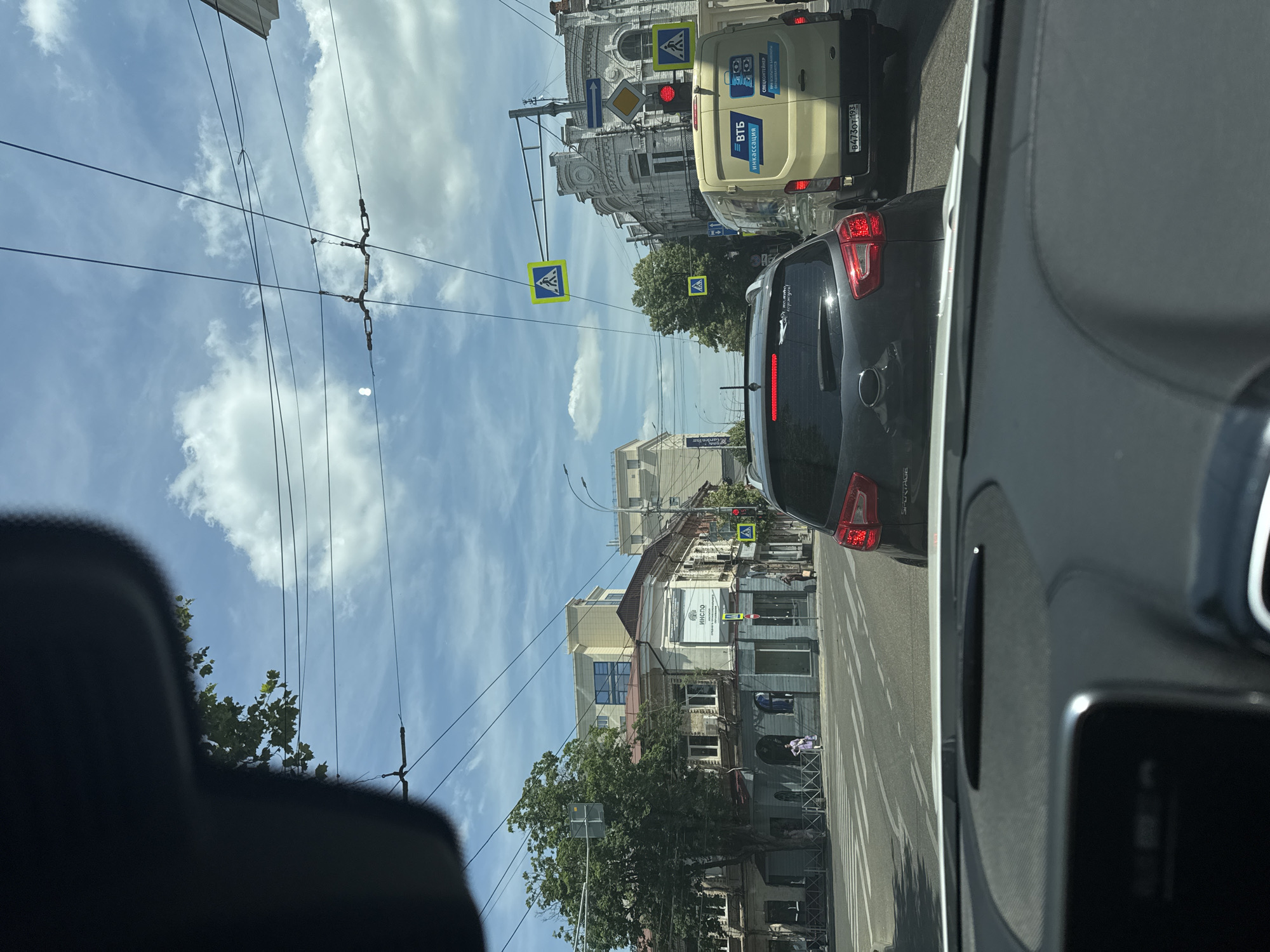 The image shows a street scene in Krasnodar, Russia, viewed from inside a vehicle. The sky is blue with scattered white clouds, suggesting daytime with fair weather. Overhead, a network of power lines crisscrosses the frame, supported by poles and structures on the buildings.
The street is lined with buildings of varying architectural styles. On the left, there are older buildings with decorative facades, some appearing to be commercial establishments. On the right, modern buildings are also visible. Several pedestrian crossing signs (yellow squares with a blue silhouette of a person walking) are posted, indicating marked crosswalks.
Traffic is present, with several vehicles visible. In the foreground on the right, part of the interior of a car, including a dashboard and part of a window, is visible. In the middle ground, a light-colored van with markings that include "ВТБ" (VTБ) and "инкассация" (cash collection) is stopped in traffic. Behind it, a dark-colored SUV is also visible. Red traffic lights are illuminated at an intersection.
A large green tree with lush foliage stands on the left side of the street, its branches extending towards the sky. Visible text includes street signs, the branding on the van, and some lettering on building signs, such as "ИНСГО" (INSGO). The overall impression is of a typical urban street scene during the day, with normal city activity and traffic.
