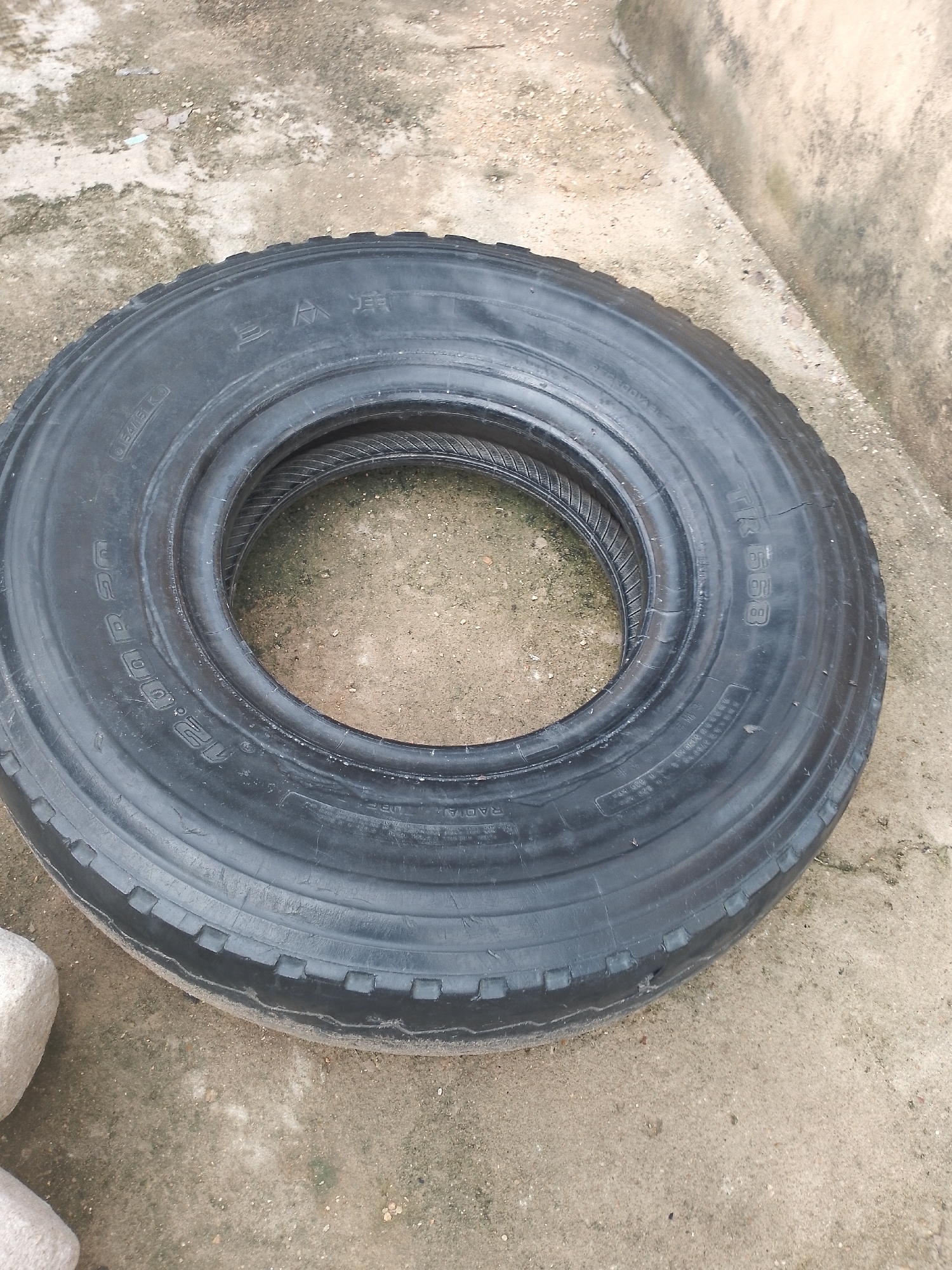 A black tire rests on a concrete surface, possibly an outdoor area. The tire is large and appears to be for a truck or heavy vehicle. There is another tire inside it, partially visible. Markings on the tire include Chinese characters and the text "12.00R20". The background shows a rough, mottled concrete ground and a slightly lighter colored concrete wall or structure. Two light-colored stones are visible in the lower left corner of the image. The lighting suggests it is daytime.
