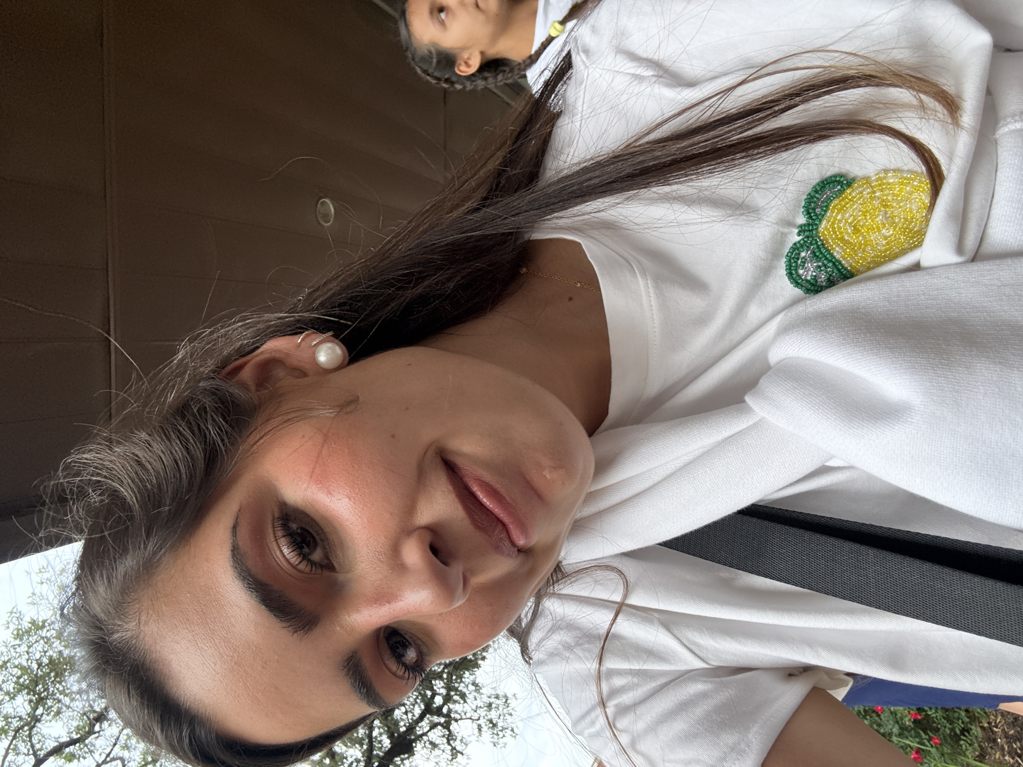 A selfie shot depicts a woman wearing a white sweater with a beaded lemon patch on the shoulder. She is wearing a pearl earring and a gold necklace. Another person is partially visible in the background, wearing a white sweater and their hair is braided with yellow accents. The location appears to be outdoors with trees visible in the background. Based on the style of clothing and general surroundings, it is plausible that this image was taken in San Antonio, Texas, United States, but there is no definitive proof in the image itself.
