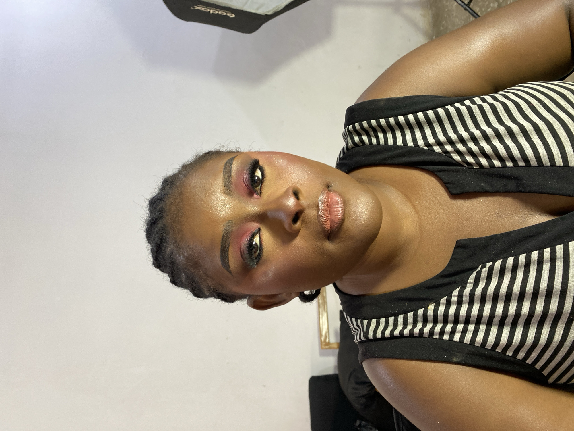 A woman with dark skin, wearing a black and white striped top, is the subject. Her hair is styled in braids. She is wearing makeup, including red eye shadow and lip color. The background is a plain white wall with a black light fixture visible in the upper left corner. The location is difficult to ascertain specifically within Gura Top, Nigeria, given the studio-like setting.