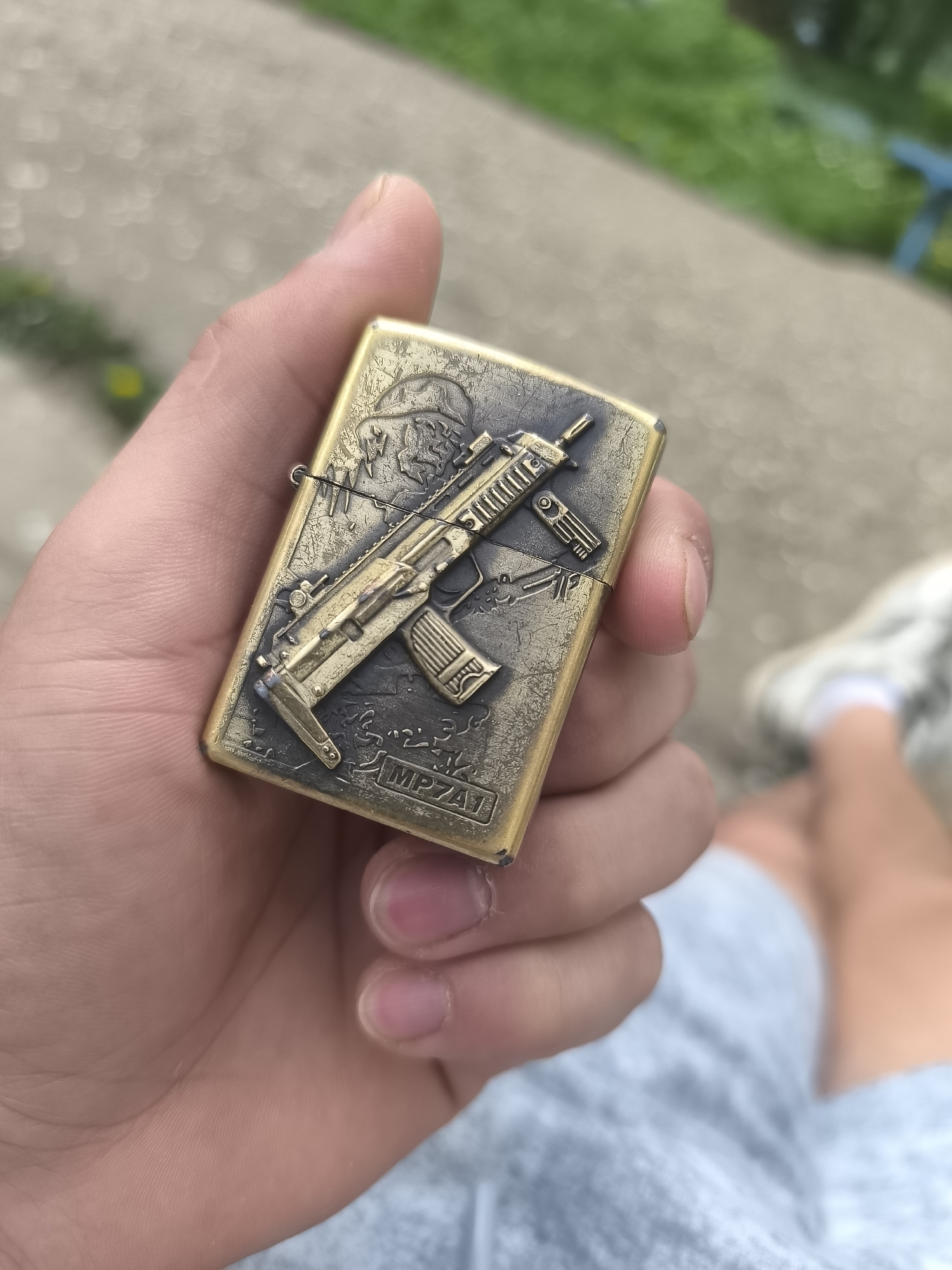 A hand holds a metal lighter that has an MP7A1 imprinted on its front in an outdoor setting.