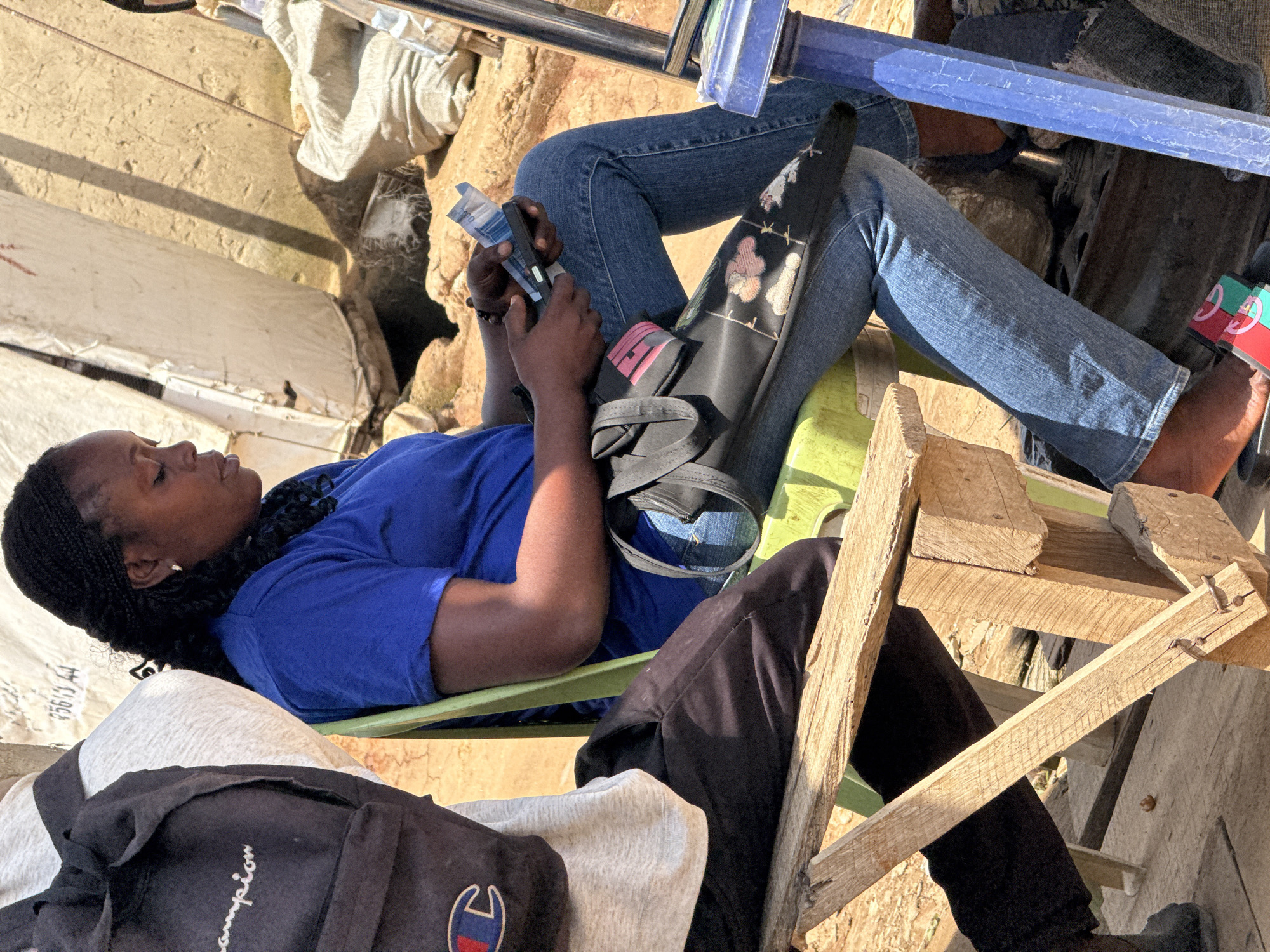 In a street scene in Abuja, Nigeria, a woman with braided hair, wearing a blue t-shirt and blue jeans, sits on a green plastic chair. She is holding a black device in her right hand and banknotes in her left hand, appearing to be engaged in a transaction or counting money. Beside her, a person wearing a grey t-shirt and black pants with a black backpack featuring the "Champion" logo is partially visible, also seated. The background consists of sandy terrain, a white tarp or tent structure, and rough textures suggesting an outdoor market or informal settlement. A wooden bench is in the foreground, and a blue-painted metal structure stands to the right of the woman.